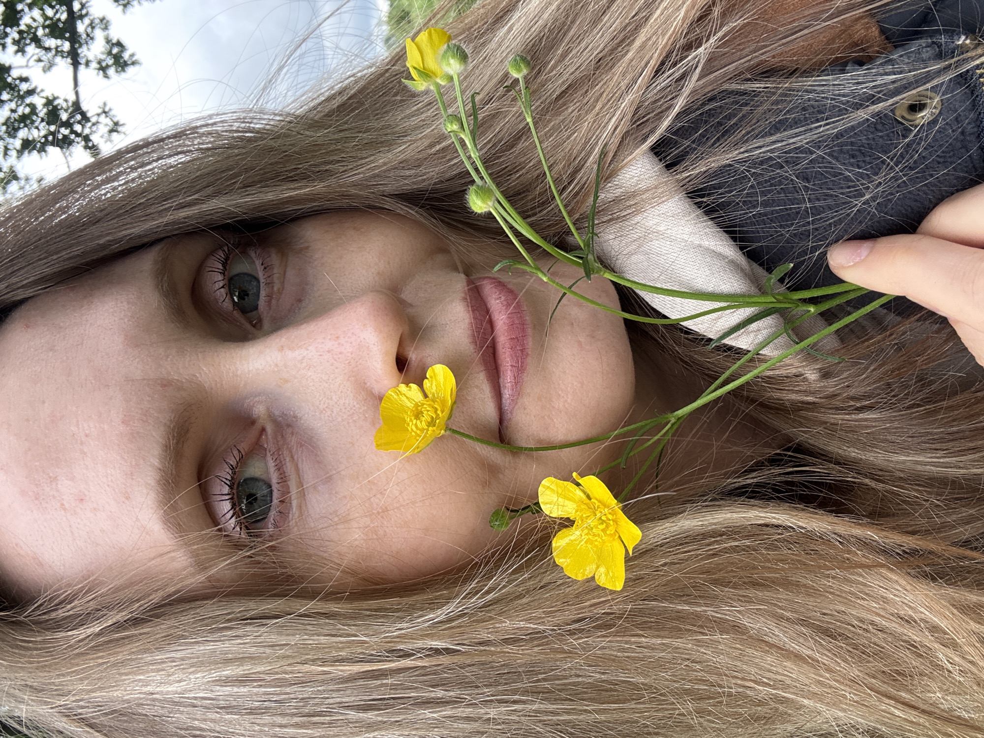 The image is a close-up, chest-up portrait of an adult female positioned outdoors. She has long, straight blonde hair that frames her face, with strands falling across her forehead and near her eyes. Her light blue eyes are open and directed towards the camera, and her lips are slightly parted in what appears to be a subtle smile. She holds a small cluster of bright yellow flowers, consistent with buttercups, against the left side of her face. One flower is positioned near her nose, and another is visible below her left eye. Her right hand, with manicured fingernails, is visible at the bottom of the frame, holding the green stems of the flowers. She wears layered clothing, including a visible cream or light-colored hooded collar over a dark, textured garment, possibly a jacket, which features a brass eyelet. The background is blurred, depicting a cloudy sky with some dark green tree foliage visible against it in the upper right. A distant, blurred green landscape suggests an open natural environment. The specific geographical location, including the city of Noviy, Russia, cannot be determined from the visual information present in the image.
