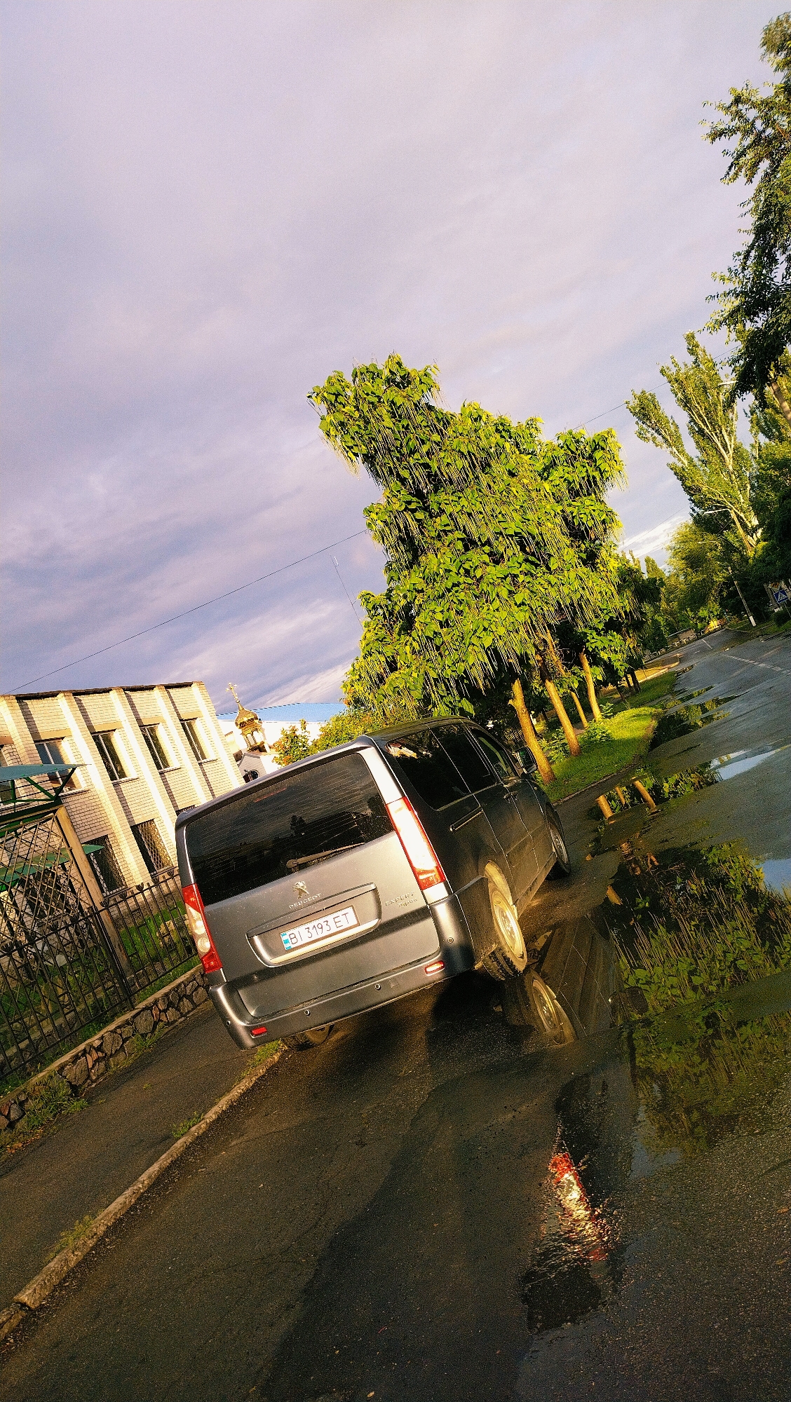 The media file shows a grey Peugeot Expert van parked on a wet asphalt road. The van is facing away from the camera, and its license plate reads "BI 393 ET". In the background, there is a modern brick building with several windows. A metal fence runs along the side of the road in front of the building.
Several trees with lush green foliage are visible on the right side of the road, casting reflections on the wet surface. The sky is overcast with streaks of purple and grey clouds, suggesting it might have rained recently or is about to rain. The lighting indicates it is either early morning or late afternoon, given the soft, warm glow on the building and the sky. The presence of puddles on the road confirms the wet weather. The setting appears to be a semi-urban or suburban area in Horishni Plavni, Ukraine, as indicated by the architectural style of the building and the road infrastructure. There are no people visible in the scene. The overall mood is serene, with the reflections in the puddles adding a touch of artistic beauty.