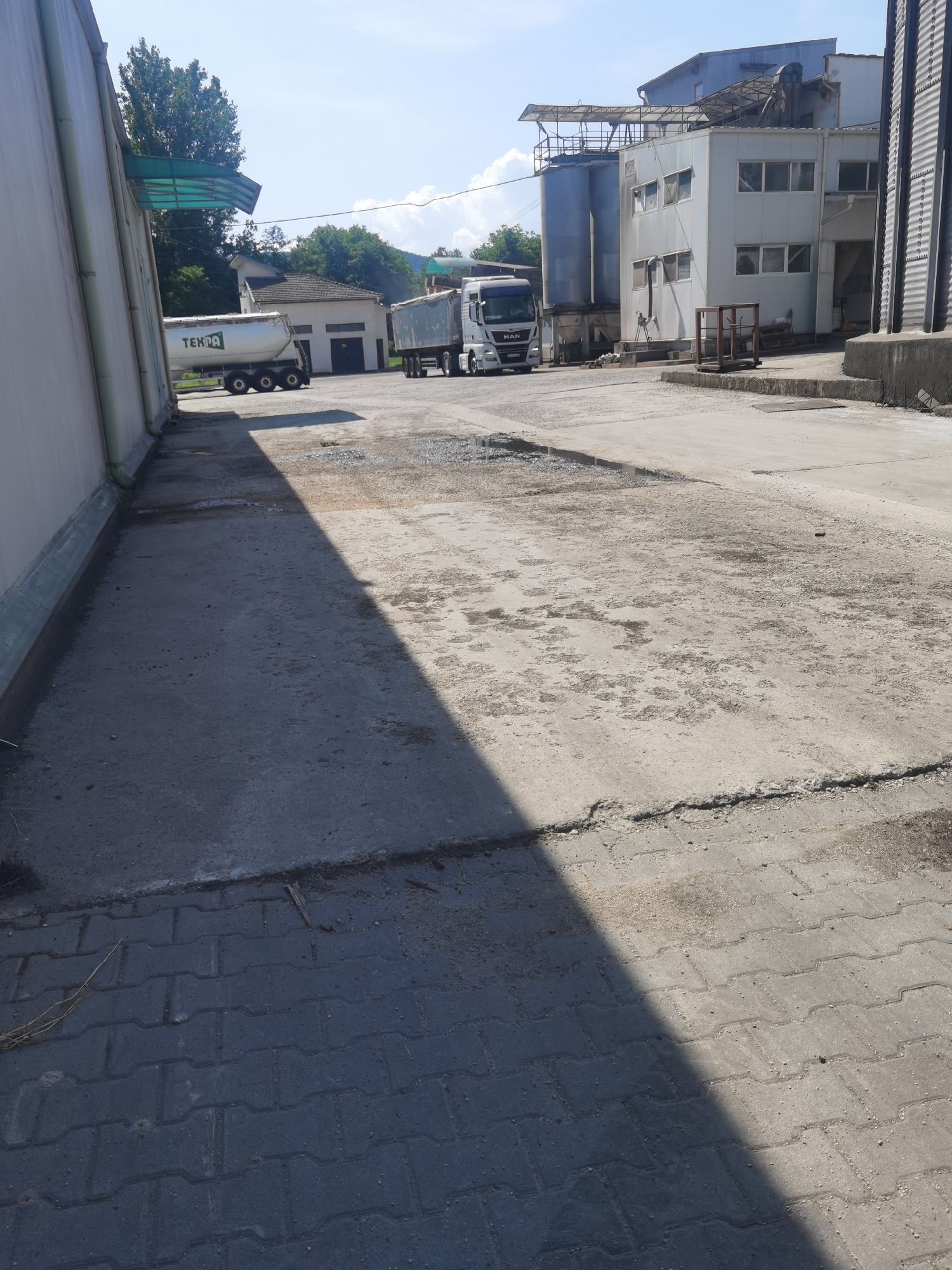 The image depicts an outdoor industrial area, likely a facility for storing or processing bulk materials, situated in Lomets, Bulgaria. In the foreground, a paved area with interlocking concrete blocks transitions to a rougher concrete surface. A significant shadow from an unseen object stretches across the pavement.
To the left, a large, light-colored building with a green awning extends along the side. Further into the scene, two white trucks are parked. The truck on the left is a tanker truck with the logo "TEKPA" visible on its side. The truck on the right appears to be a standard tractor-trailer.
In the background, a multi-story industrial building with a light-colored facade and windows is visible, adjacent to large cylindrical storage tanks. The sky is bright blue with some scattered clouds, suggesting a sunny day. The environment appears functional and utilitarian, typical of an industrial or agricultural processing site. No people are visible in the image.