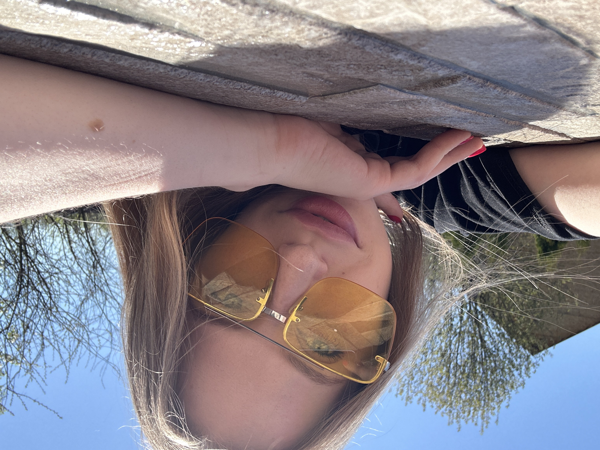 A woman with light brown hair wearing yellow-tinted sunglasses is reclining, with a portion of her left arm visible with a mole, touching a gray tiled surface. Her right hand, with red nail polish, also rests on the surface. She is wearing a black top. The background shows trees and a blue sky, suggesting an outdoor location, potentially in Chaikovsky, Russia. The photo is taken from a low angle.