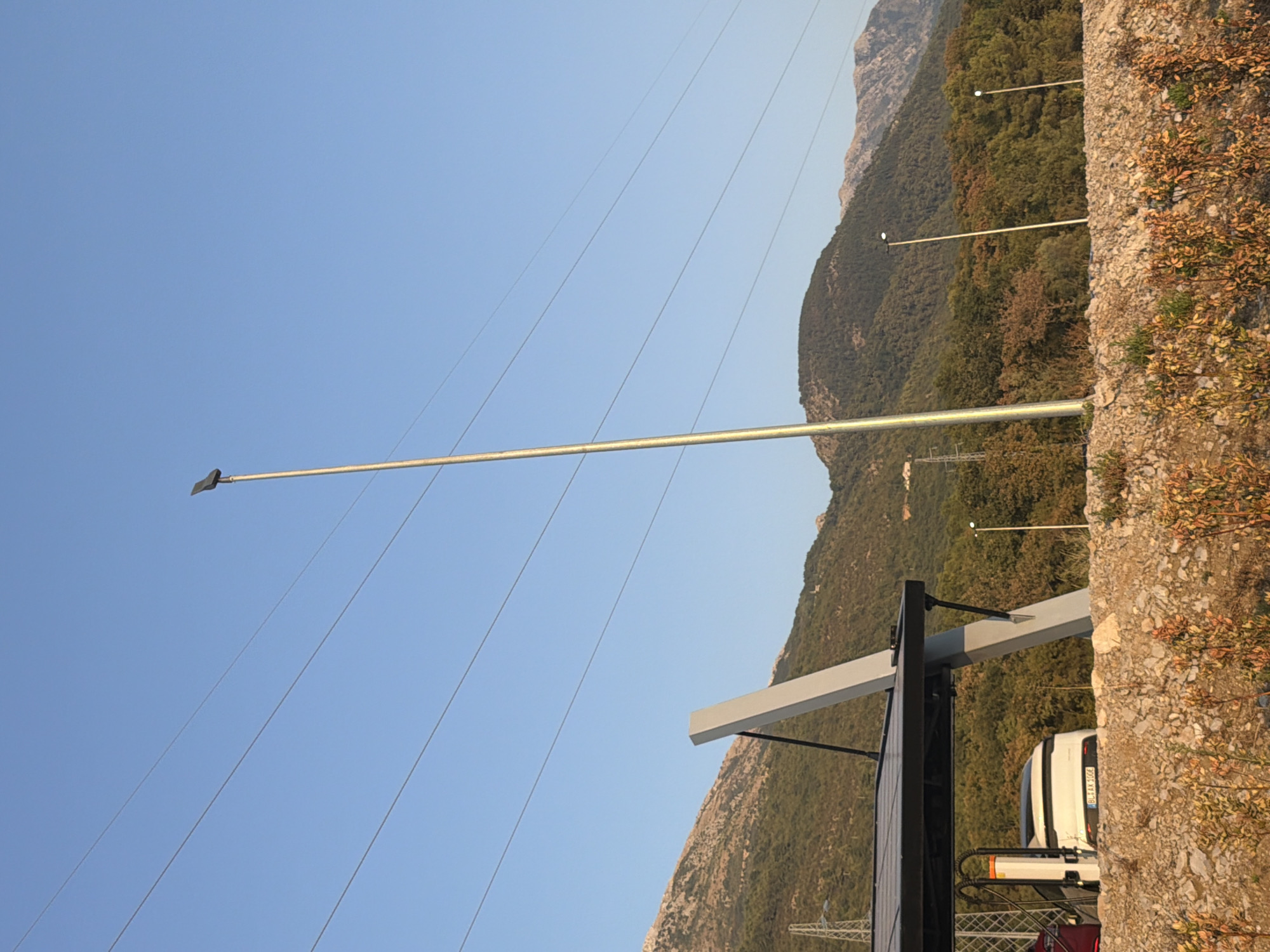 The image captures a scenic view of Dub, Montenegro, featuring an electric vehicle charging station with a white car plugged in. The charging station is equipped with solar panels, suggesting a sustainable energy source. In the background, lush green mountains rise against a clear blue sky, with power lines stretching across the frame. A tall pole with a light fixture stands prominently in the foreground, adding to the urban infrastructure amidst the natural beauty. The ground is covered with gravel and scattered vegetation, indicating a rural or semi-rural setting.