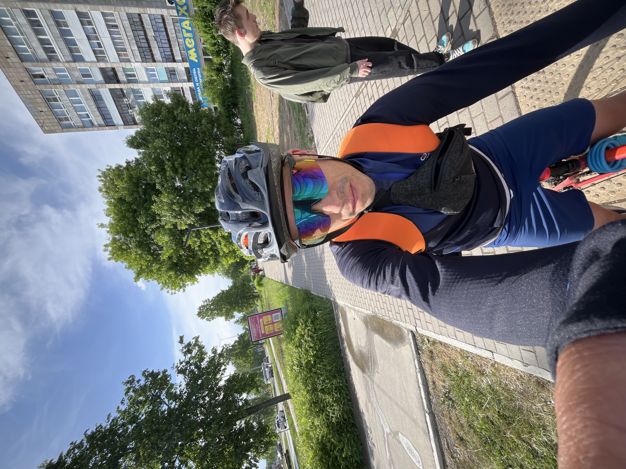 The image depicts a person on a bicycle, wearing a helmet and reflective sunglasses, taking a selfie. The individual is wearing cycling attire consisting of a navy blue top with an orange reflective vest and dark blue shorts. A small black bag is strapped around their waist. Another individual, a young man in a green jacket and dark pants, walks on the sidewalk beside the cyclist.
The scene is set on a sidewalk in the city of Chaikovsky, Russia. A tall apartment building is visible in the background, featuring multiple windows. The building has an advertisement in Russian. Trees line the street and sidewalks. A sign on the side of the road advertises Faberlic. Cars and other vegetation are visible along the roadside. The sky is visible above the trees, showing a partly cloudy, sunny day.