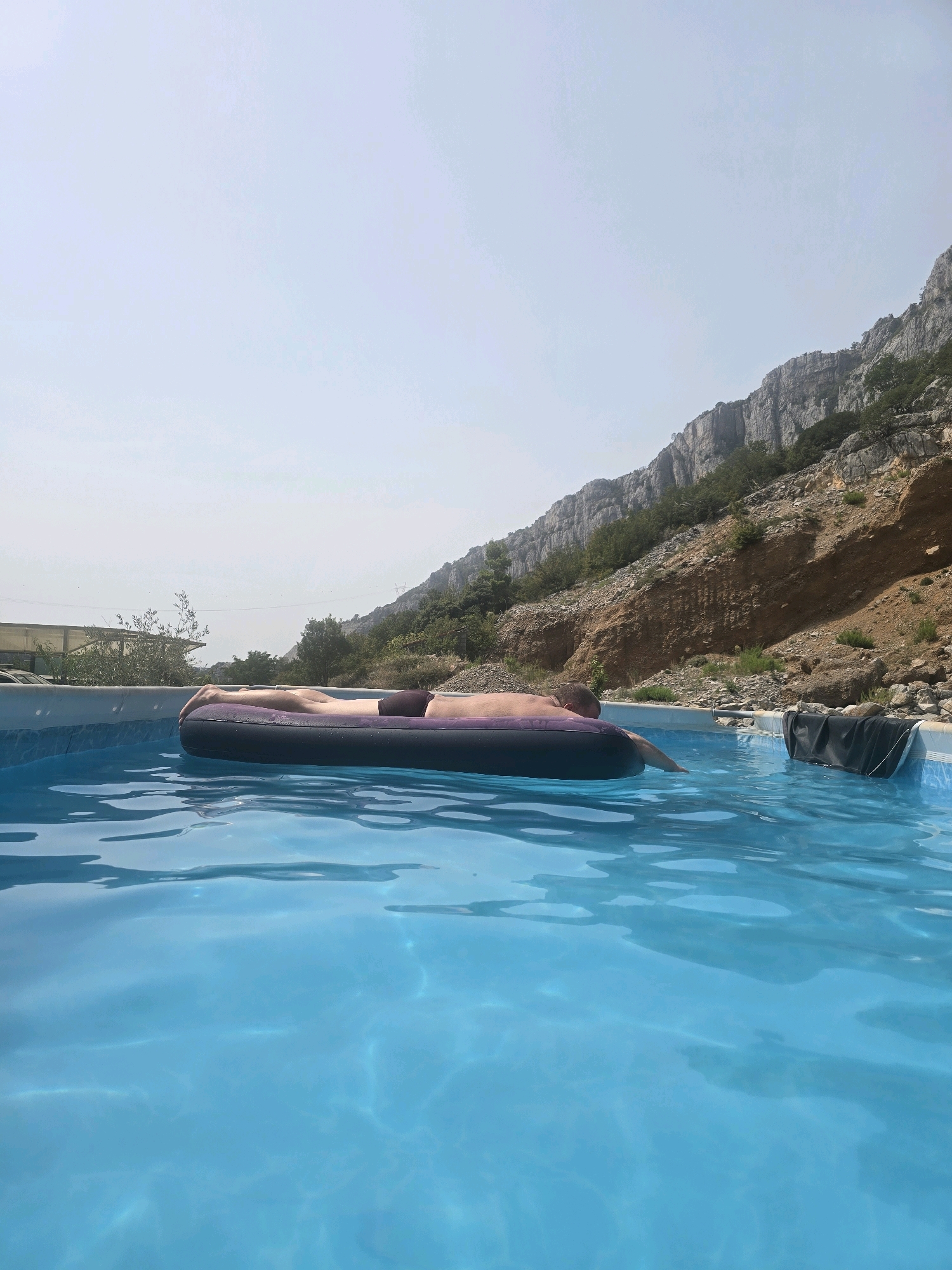 In Podgorica, Montenegro, a person is resting on a purple inflatable raft in a bright blue swimming pool. The pool is surrounded by a white rim. In the background, a steep, rocky mountainside with sparse vegetation rises towards a hazy sky. To the left of the mountainside, a building with a light-colored roof and windows is visible, along with some trees and a parked vehicle. The sunlight casts ripples on the surface of the water.