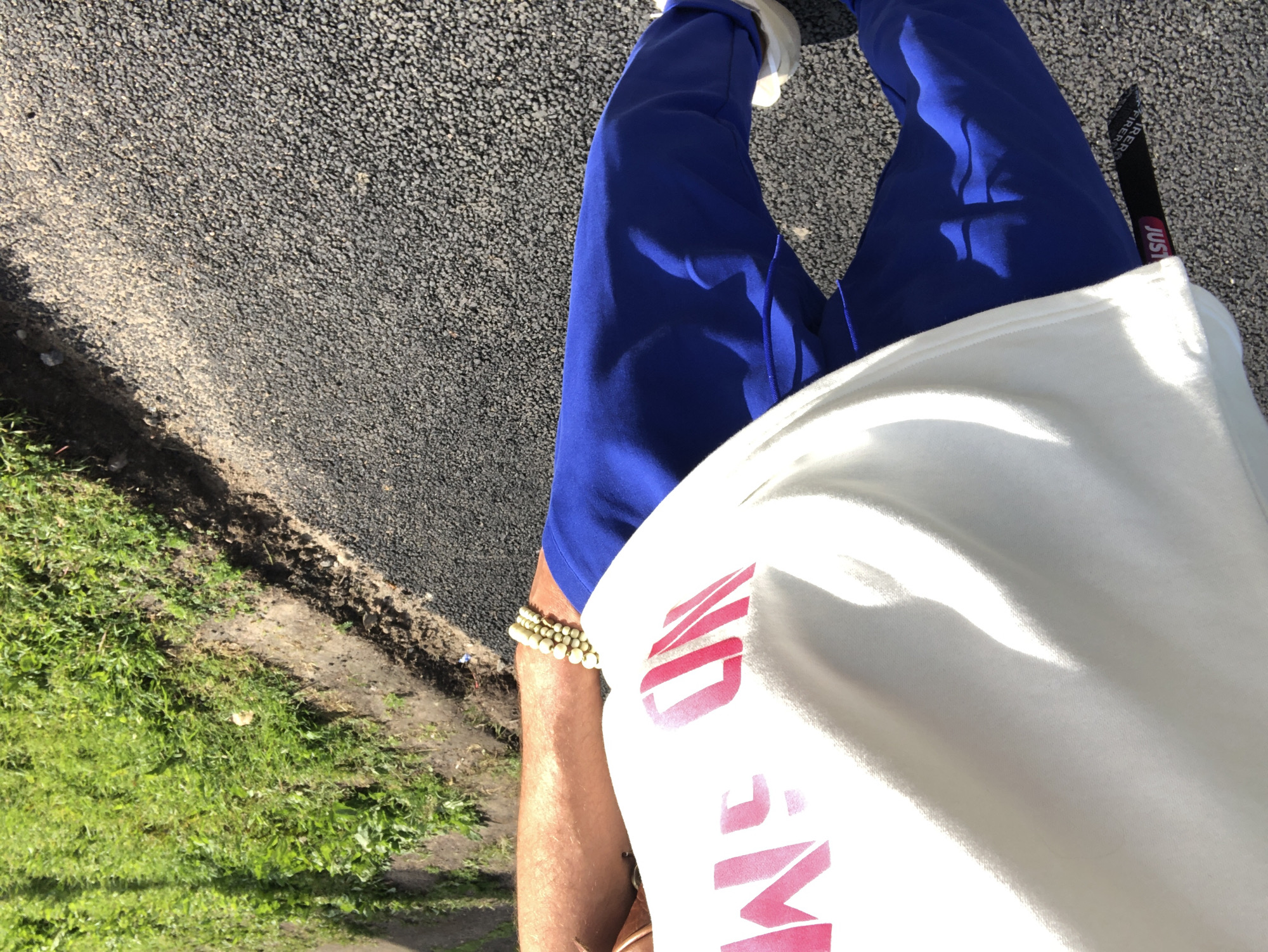 This is a first-person perspective photo of a person's legs and torso, taken outdoors. The person is wearing bright blue athletic pants and a white t-shirt with pink lettering. The t-shirt has a large, bold pink graphic design that appears to be text, but only parts of the letters are visible. A black tag with white lettering, possibly a brand label, is tucked into the waistband of the pants on the right side. On the left wrist, a beaded bracelet is visible.
The setting appears to be a paved path or road next to a grassy area. The ground is dark gray and textured, consistent with asphalt or a similar surface. The grass is green and appears to be well-maintained. The lighting suggests it is daytime, with shadows cast by the person, indicating the sun is out. There are no clear indications of the specific location within Saint Petersburg, Russia, other than the provided context. The overall mood is casual and active, likely depicting someone walking or standing outdoors.