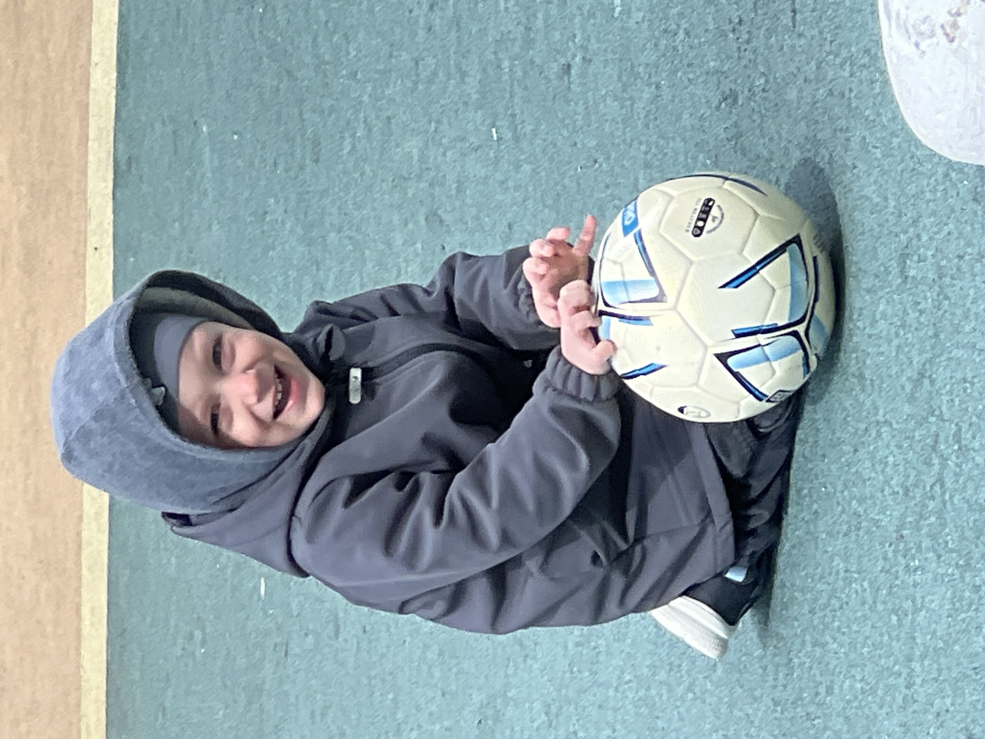 A young child wearing a gray hooded jacket and a gray beanie is holding a soccer ball. The ball is white with blue accents. The child is smiling and appears to be sitting on a surface with a light blue-green textured pattern. A strip of light brown or tan flooring is visible along the left edge of the image. Part of a white object is visible in the upper-right corner. The location is specified as Mytishchi, Russia.