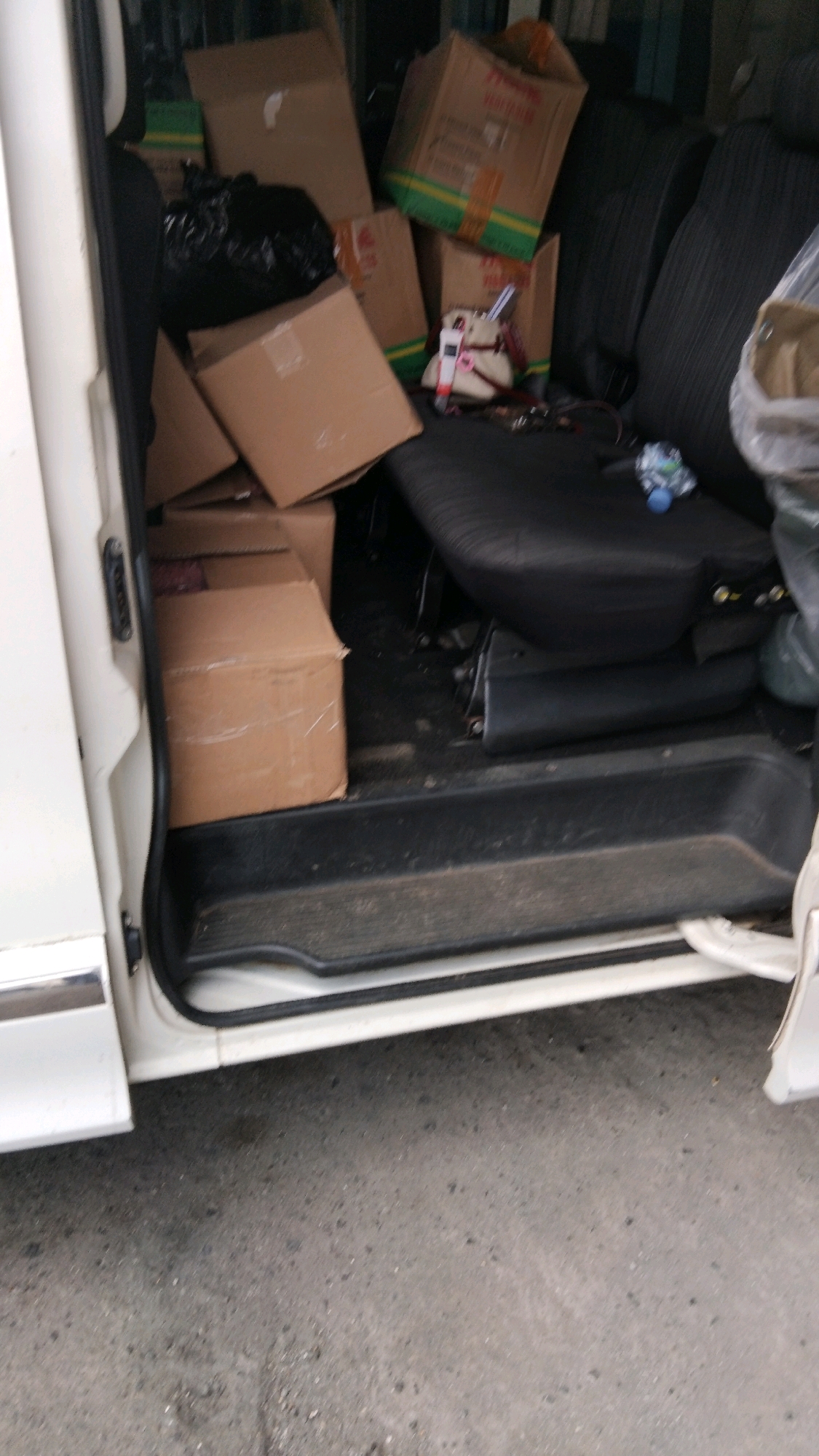 The image shows the interior of a white van, packed with cardboard boxes of various sizes. The boxes are stacked haphazardly, with some in the front passenger area and others on the floor. A black garbage bag is also visible among the boxes. The van's doors are open, revealing the interior. The scene appears to be a typical street in Pekalongan, Indonesia, with a concrete surface visible on the right side of the image. There are no people visible in the image.