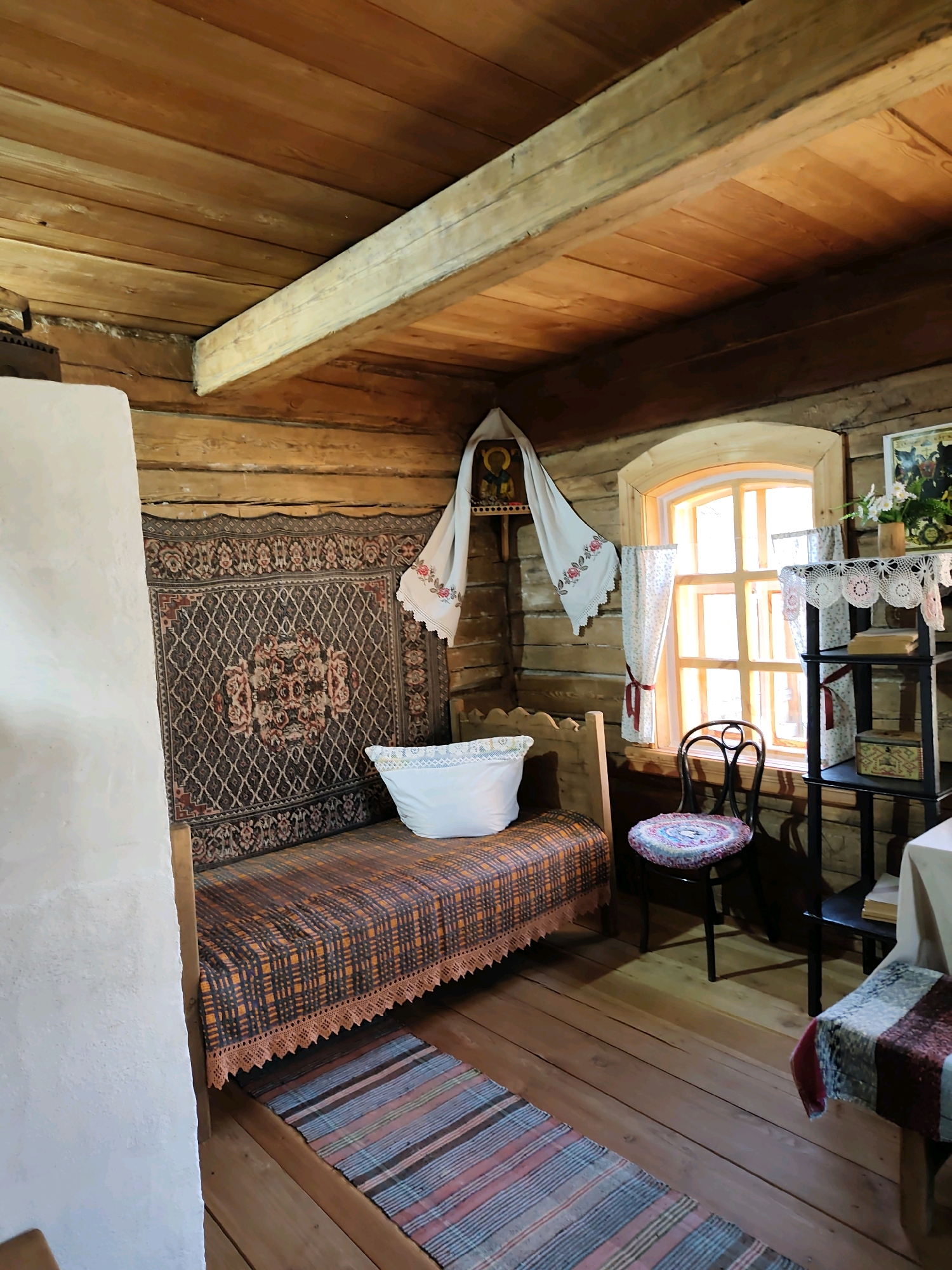 The image depicts the interior of a traditional wooden house, likely a museum exhibit in the city of Taltsy, Irkutsk Oblast, Russia. The room is furnished in a rustic style, featuring log walls and a wooden plank ceiling supported by visible beams.
Dominating the scene is a simple wooden bed, covered with a patterned blanket and a white pillow. A large, intricately patterned rug hangs on the wall above the bed, and a smaller striped rug lies on the wooden floor in front of it. To the right of the bed, a dark wooden chair with a colorful cushion stands near a window. The window itself is divided into multiple panes and is adorned with lace curtains.
On a shelf to the right of the window, there are books, a decorative box, and a vase with flowers. A white fabric, possibly a towel or shroud with embroidered flowers, hangs on the wall to the left of the window, with an icon above it. A white-tiled stove is partially visible on the far left of the image. The floor is made of worn wooden planks, adding to the authentic, aged feel of the space. The overall impression is one of a preserved historical dwelling, showcasing traditional Russian rural life.