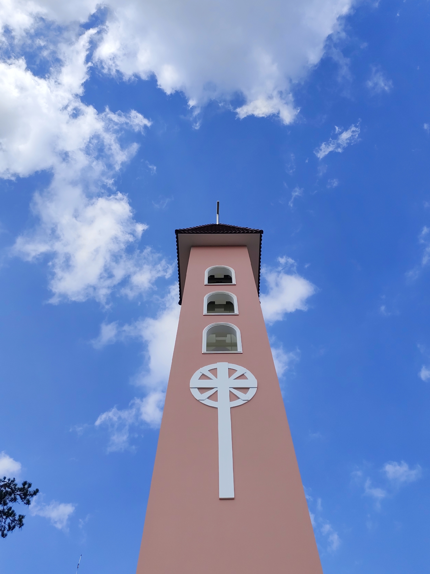 A tall, pink tower with a white cross on its facade and three arched windows near the top is shown against a blue sky with scattered white clouds. A small black cross is positioned atop the tower, which has a brown roof. The image is a low-angle shot emphasizing the height of the structure. The location is Đa Thọ, Vietnam.