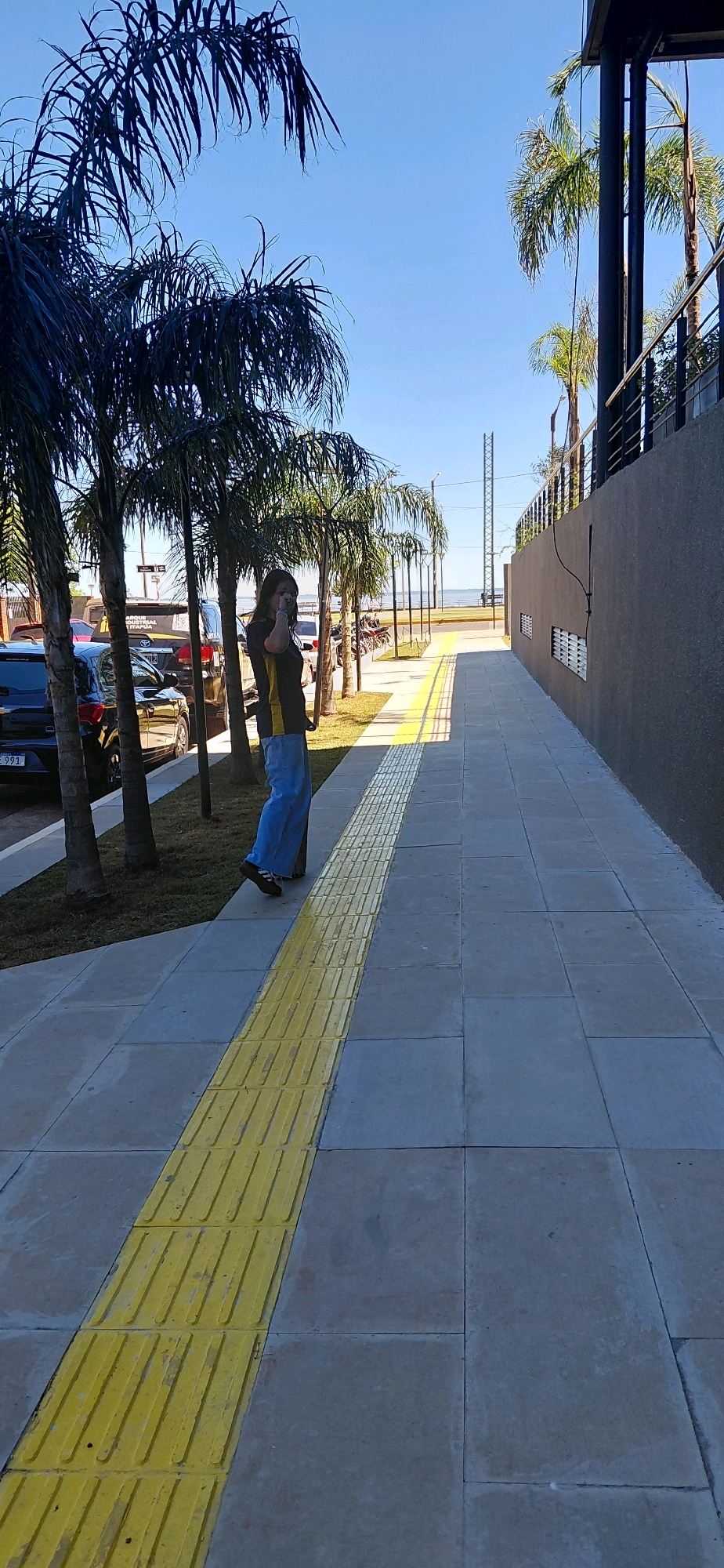 This media file depicts a sunny day in Encarnación, Paraguay. The scene is a paved walkway beside a grassy area with several palm trees. A woman is walking along the sidewalk, looking down and touching her face, possibly speaking on a phone or adjusting a mask. She is wearing a dark t-shirt with a yellow stripe and light blue jeans. The walkway has a tactile paving strip with raised studs, common for visually impaired pedestrians.
In the background, parked cars are visible, including a dark SUV with a license plate ending in "991". Beyond the cars and trees, a body of water, likely the Paraná River, is visible along with a distant horizon and a clear blue sky. The architecture of a building with dark walls and a metal railing is on the right side of the walkway. A tall metal tower structure can be seen in the distance, as well as some other people and motorcycles further down the waterfront.
The time of day is daytime, suggested by the bright sunlight casting shadows. The weather appears clear and warm. The location cues, such as the palm trees, waterfront, and the implied tropical or subtropical climate, along with the explicit mention of Encarnación, Paraguay, confirm the setting. There is some visible text on a yellow sign behind the parked cars, but it is not legible.