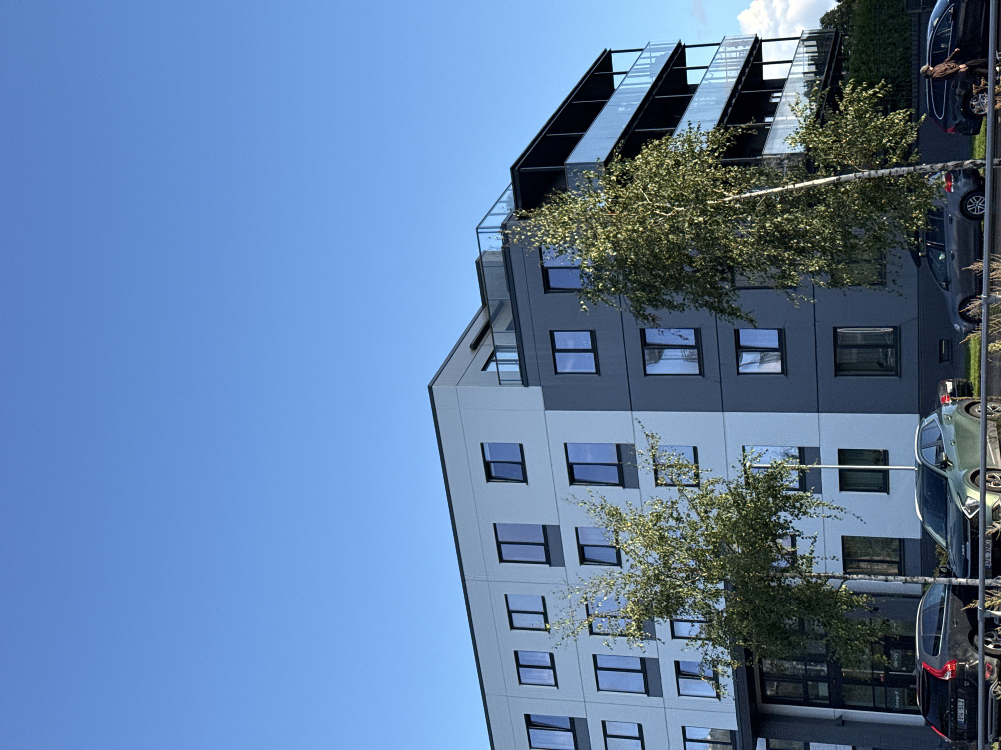 In the city of Tallinn, Estonia, a modern multi-story building with light blue and dark grey facades stands under a clear blue sky. The building features numerous rectangular windows and balconies with glass railings. In front of the building, a thin birch tree with green foliage is positioned slightly to the left of center. The street level shows parked cars, including a black Volvo SUV with license plate "914 SLZ" on the left, a green SUV in the middle, and a black car on the right. A person is visible near the black car on the right, appearing to open the driver's side door. The environment suggests an urban setting with a clear day.