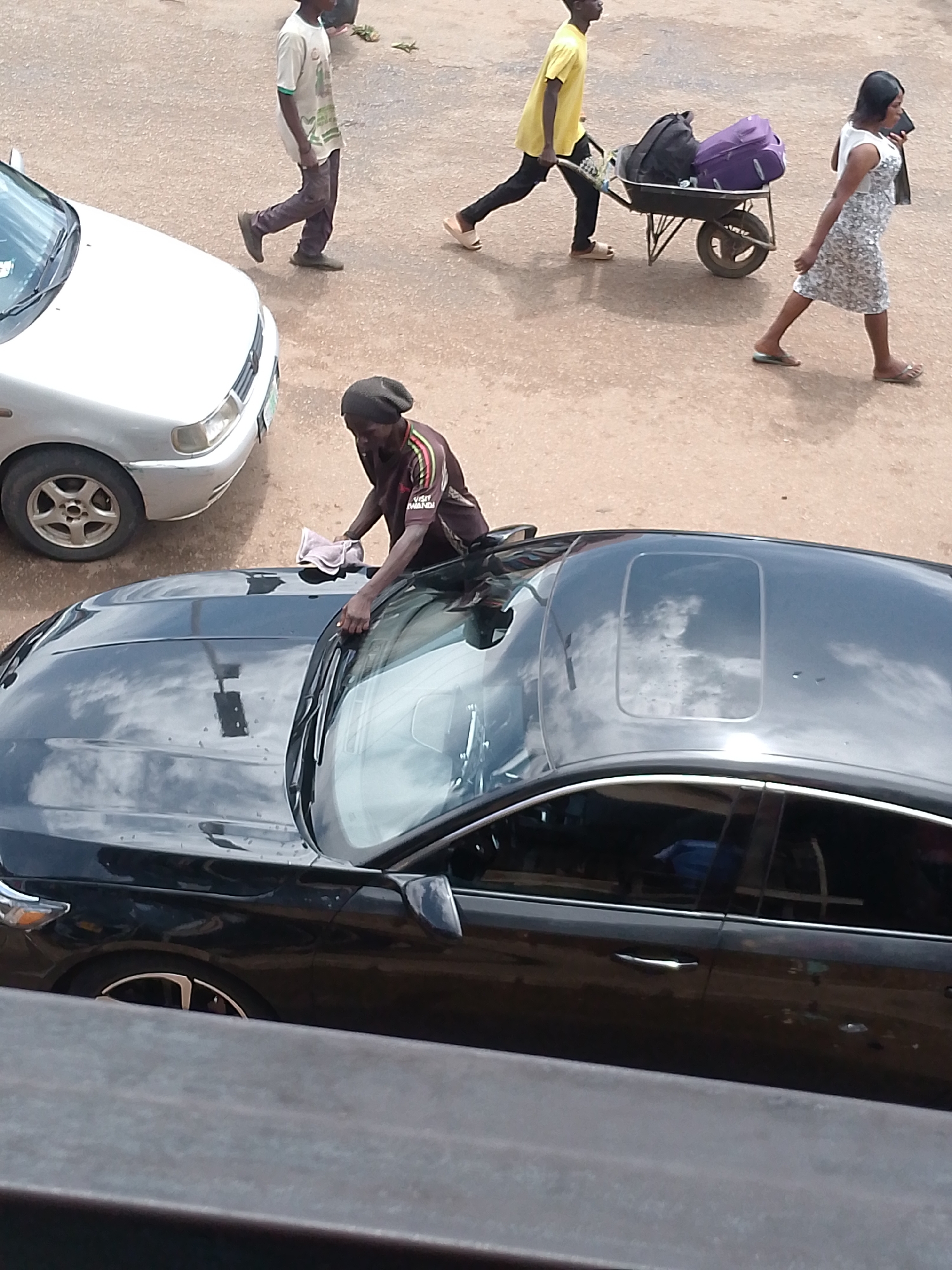 In the city of Bukuru, Nigeria, a man wearing a dark beanie and a brown shirt with colorful stripes is cleaning the hood of a black car. He is holding a light pink cloth in his hands and appears to be wiping the car. To the left of the black car, a silver sedan is parked. In the background, a person in a white t-shirt and dark pants walks past, followed by a person in a yellow shirt pushing a wheelbarrow loaded with two bags, one purple and one black. A woman in a white dress with a floral pattern walks behind them, holding a black object, possibly a phone, in her right hand. The ground is paved with a dusty, reddish-brown surface. The scene is captured from an elevated perspective, suggesting it was taken from a higher vantage point.