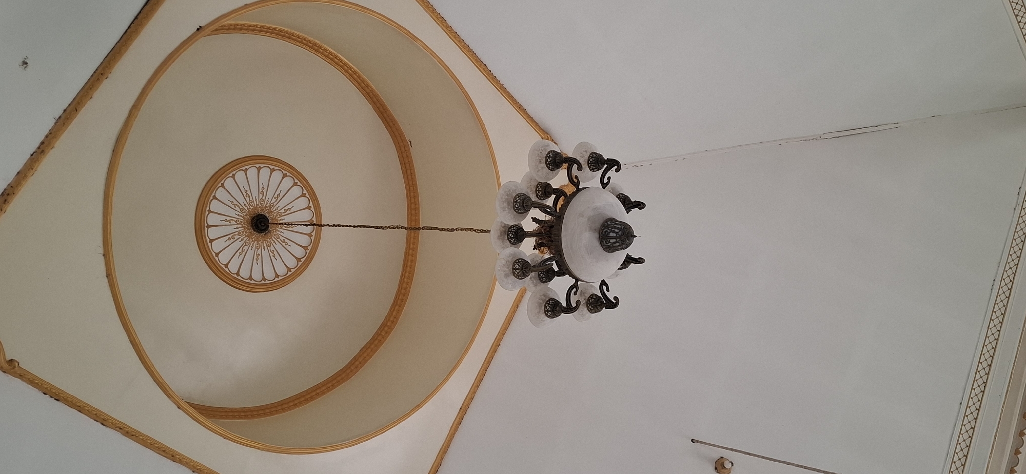 A chandelier hangs from a decorative ceiling in what appears to be a building in Wonorejo, Indonesia. The ceiling features concentric gold trim and a central circular medallion with an ornate design. The chandelier itself is made of dark metal with multiple frosted glass globes. The surrounding walls are plain white.