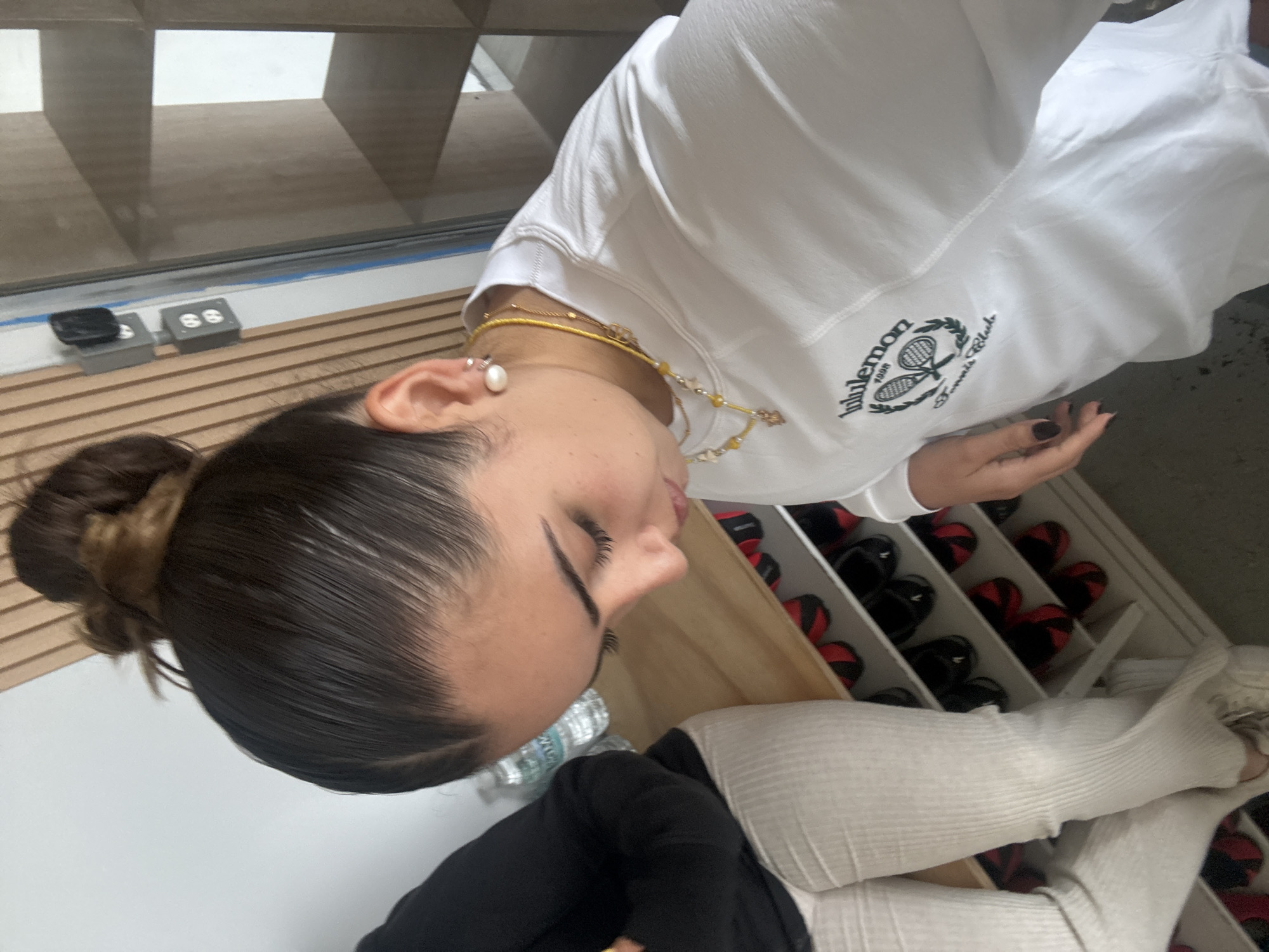 The image shows a young woman in a white sweatshirt with a "Lululemon Tennis Club" logo, wearing a layered gold necklace and pearl earrings. Her dark hair is pulled back in a bun. She is wearing ribbed white socks and dark clothing. The background shows a grid-like structure, possibly shelving or a window design, and a wooden surface. Further in the background, there's a shoe rack filled with black and red shoes. A water bottle is also visible on the table surface next to her. There is no specific information in the image that would indicate the location is Puebla, Mexico.