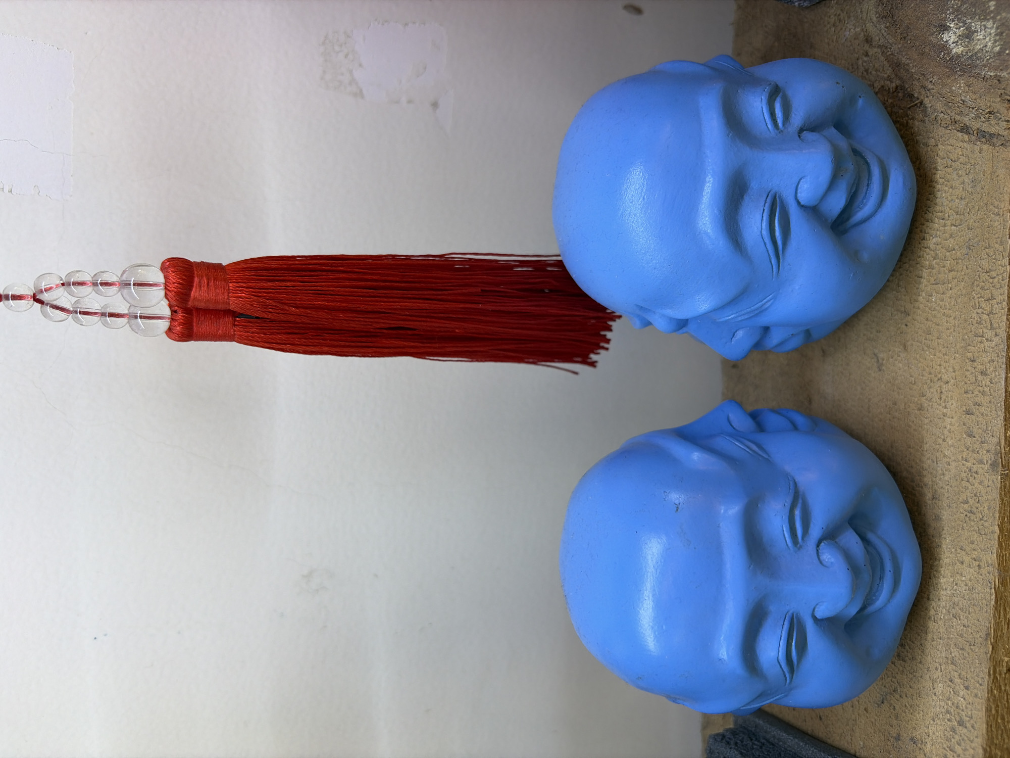 Two blue, multi-faced Buddha heads are displayed on a wooden surface. Above them, a red tassel with clear beads hangs from a string. The background is a textured white wall with some discoloration. The scene appears to be indoors, possibly in a shop or display area.