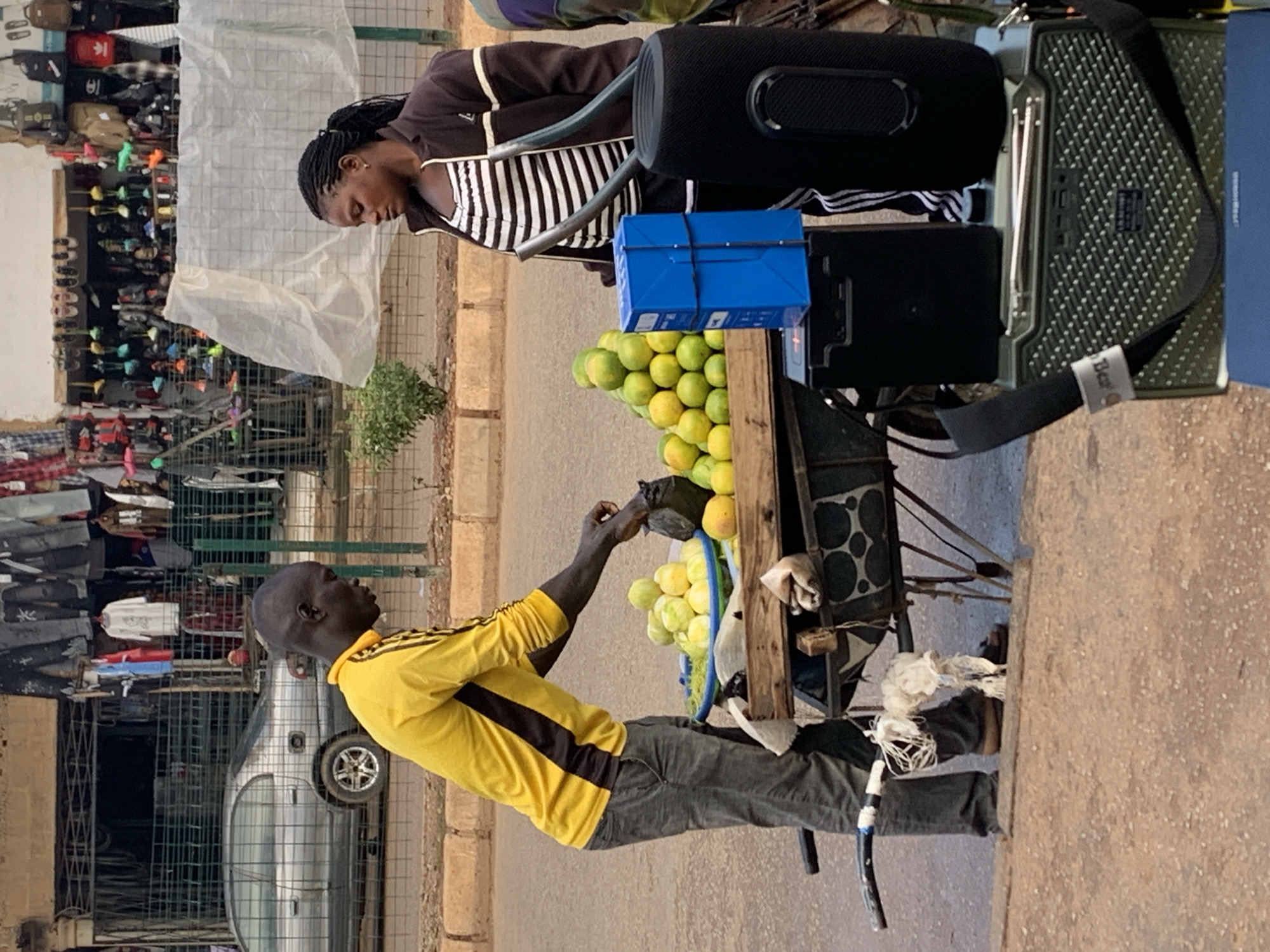 The image shows an outdoor scene in what appears to be a market setting in Bukuru, Nigeria. The primary subjects are two individuals, a woman and a man, positioned near a cart laden with produce, predominantly yellow fruits that resemble lemons or limes.
The woman, with dark braided hair, is wearing a dark jacket over a horizontally striped top. She is looking down, her expression obscured. A large black portable speaker is visible behind her.
The man, with dark skin and a shaved head, is wearing a bright yellow long-sleeved shirt with black stripes and dark gray trousers. He is actively engaged with the fruit on the cart, his hand reaching towards a cluster of the yellow fruits. His body language suggests he is either selecting or arranging the produce.
The cart itself is rustic, made of wood and metal, and is equipped with wheels. A bright blue box is placed on top of the fruit. Various other items, including what looks like packaged goods, are also on the cart. In the background, a silver car is parked, and shelves stocked with clothing and bags are visible behind a green metal fence, indicating a bustling marketplace.
The lighting suggests it is daytime, possibly late morning or afternoon, with the sun casting shadows. The ground is paved with rough material, and a low brick wall runs along the edge of the street. The overall impression is that of a busy commercial environment where daily transactions are taking place. No visible text is discernible in the image.
