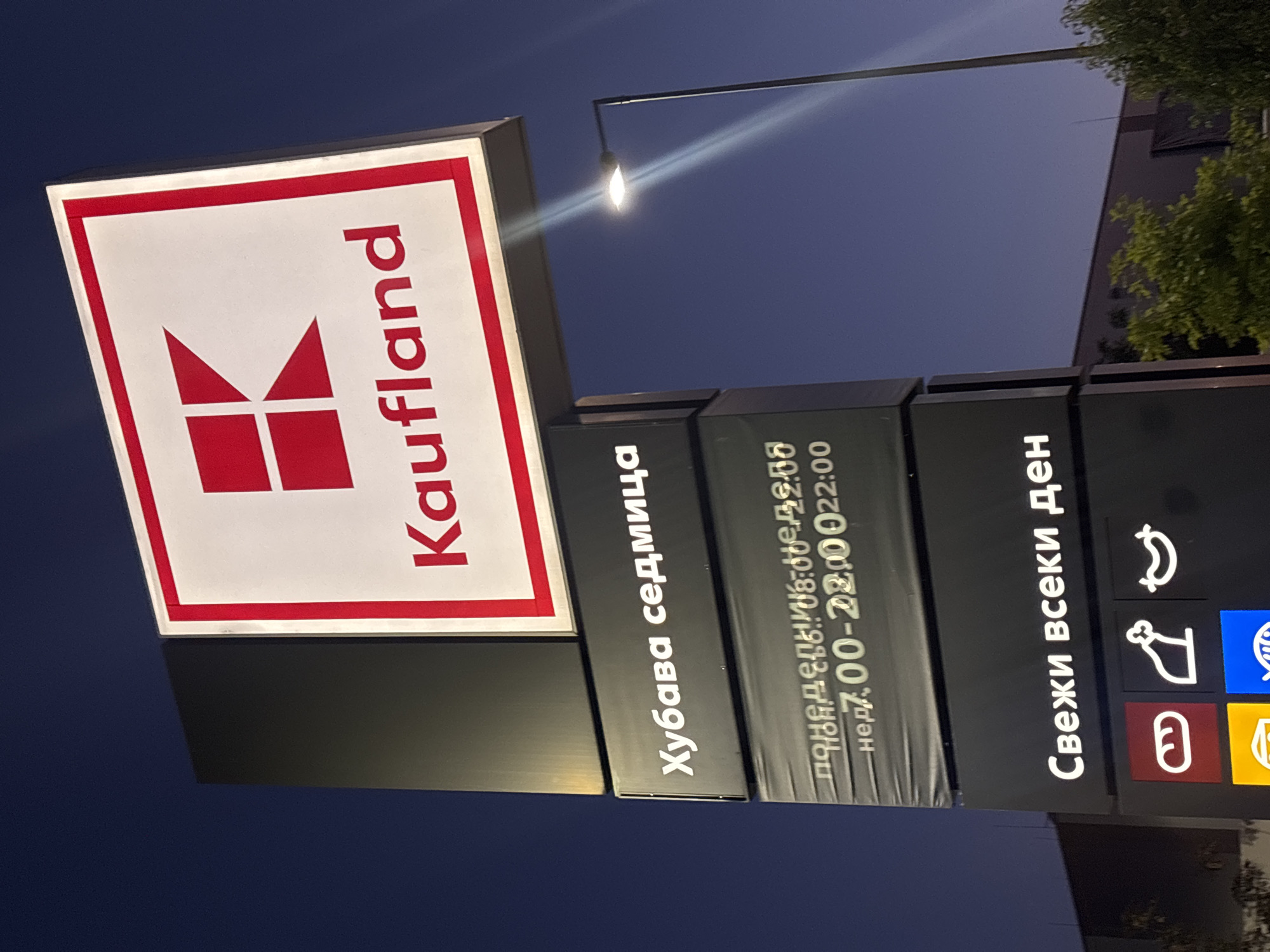 This is a nighttime, eye-level, medium shot of a lit-up sign for a Kaufland supermarket. The sign is tall and rectangular, with the Kaufland logo at the top, featuring a red "K" within a white square surrounded by a red border. Below the logo, the text "Kaufland" is written in red. Further down the sign, there is text in Cyrillic. The first line reads "Хубава седмица" (Hubava sedmitsa), meaning "Nice week". The second line appears to indicate store hours, with "понед. съб.: 08:00-22:00" (poned. sub.: 08:00-22:00), meaning "Monday-Saturday: 08:00-22:00", and "нед.: 7:00-22:00" (ned.: 7:00-22:00), meaning "Sunday: 7:00-22:00". The third line displays "Свежи всеки ден" (Svezhi vseki den), meaning "Fresh every day". Below this, there are icons representing food items: a loaf of bread, a chicken or turkey, and a sausage.
The sign is illuminated, and a streetlamp is visible to the right of the sign, casting a bright light. The sky is dark blue, indicating it is night or dusk. In the background, the facade of a building is visible, along with some trees. The overall scene suggests a commercial area in Varna, Bulgaria, likely a shopping district or a street with supermarkets. There are no people visible in the image. The image appears to have been taken from a low angle looking up at the sign.