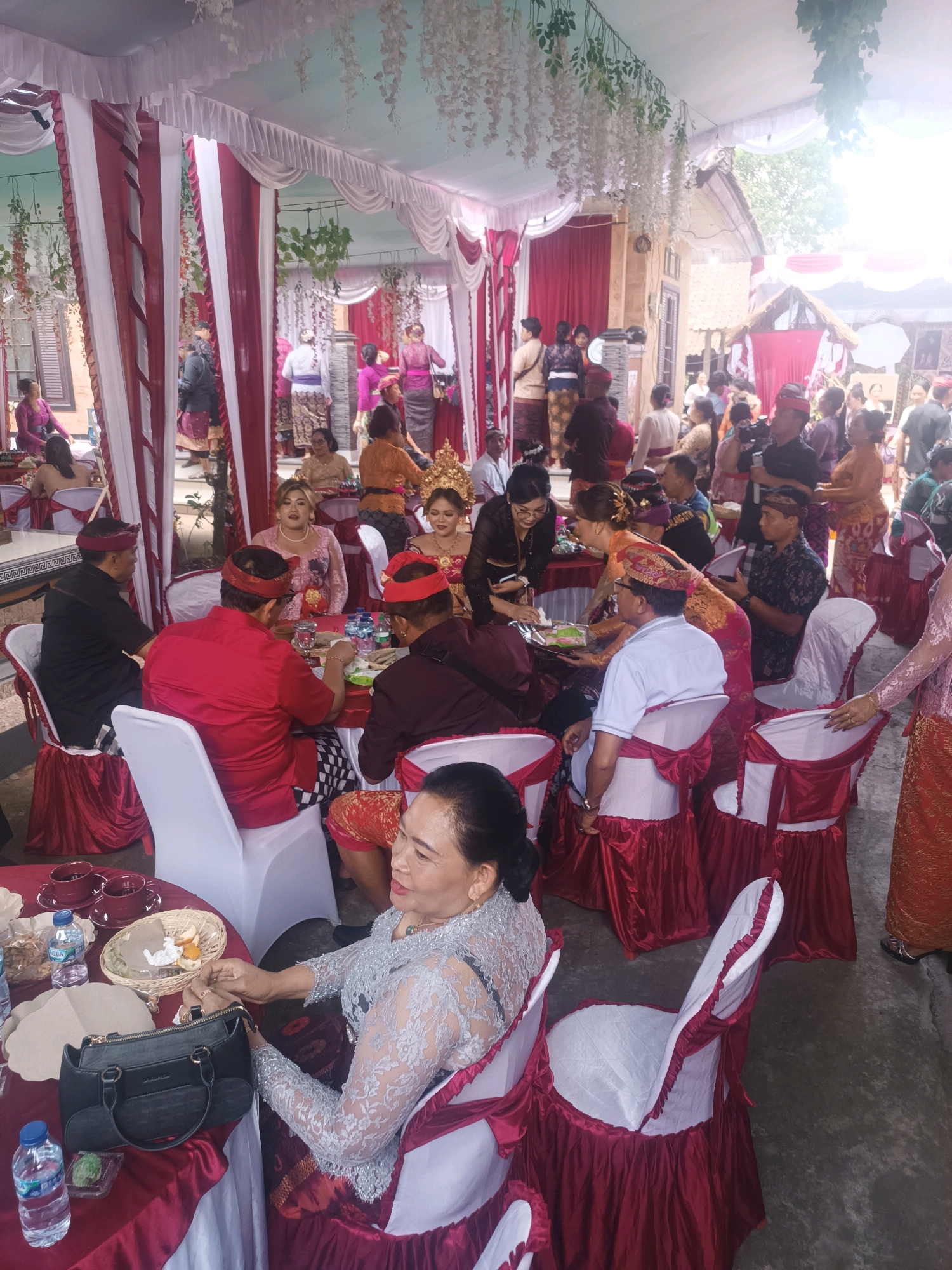 The image captures an outdoor gathering, likely a ceremony or reception, held under a canopy decorated with white fabric and hanging artificial white flowers. The event appears to be in Penebel, Indonesia, given the traditional attire and cultural elements visible. Numerous people are present, seated at tables covered with dark red tablecloths and dressed in a combination of traditional and formal wear.
In the foreground, a woman in a light blue, lace-patterned top and a dark red skirt is seated, facing the right of the frame. She has dark hair and is wearing jewelry. On the table in front of her are two bottles of Aqua water, a black handbag, and a woven basket containing what appears to be snacks or offerings. Two dark red teacups are also visible on the table.
To the left of the foreground, three men are seated at a table. The man closest to the viewer is wearing a black shirt and a red headband. The man in the center is wearing a red shirt and a red headband, with a black and white checkered pattern on his lower garment. The third man, further back, is wearing a dark maroon shirt and also has a red headband. All three are seated on white chairs covered with dark red skirting.
In the background, more guests are seated at tables, and others are standing and interacting. Several women are adorned in traditional Indonesian attire, some with elaborate headwear, suggesting a significant cultural event. There are decorative red and white drapes framing the area, and in the far background, a traditional Indonesian building or structure with a thatched roof is visible. The lighting suggests it is daytime, with sunlight filtering through the canopy. The overall scene depicts a lively and organized event with a strong sense of community and tradition.
