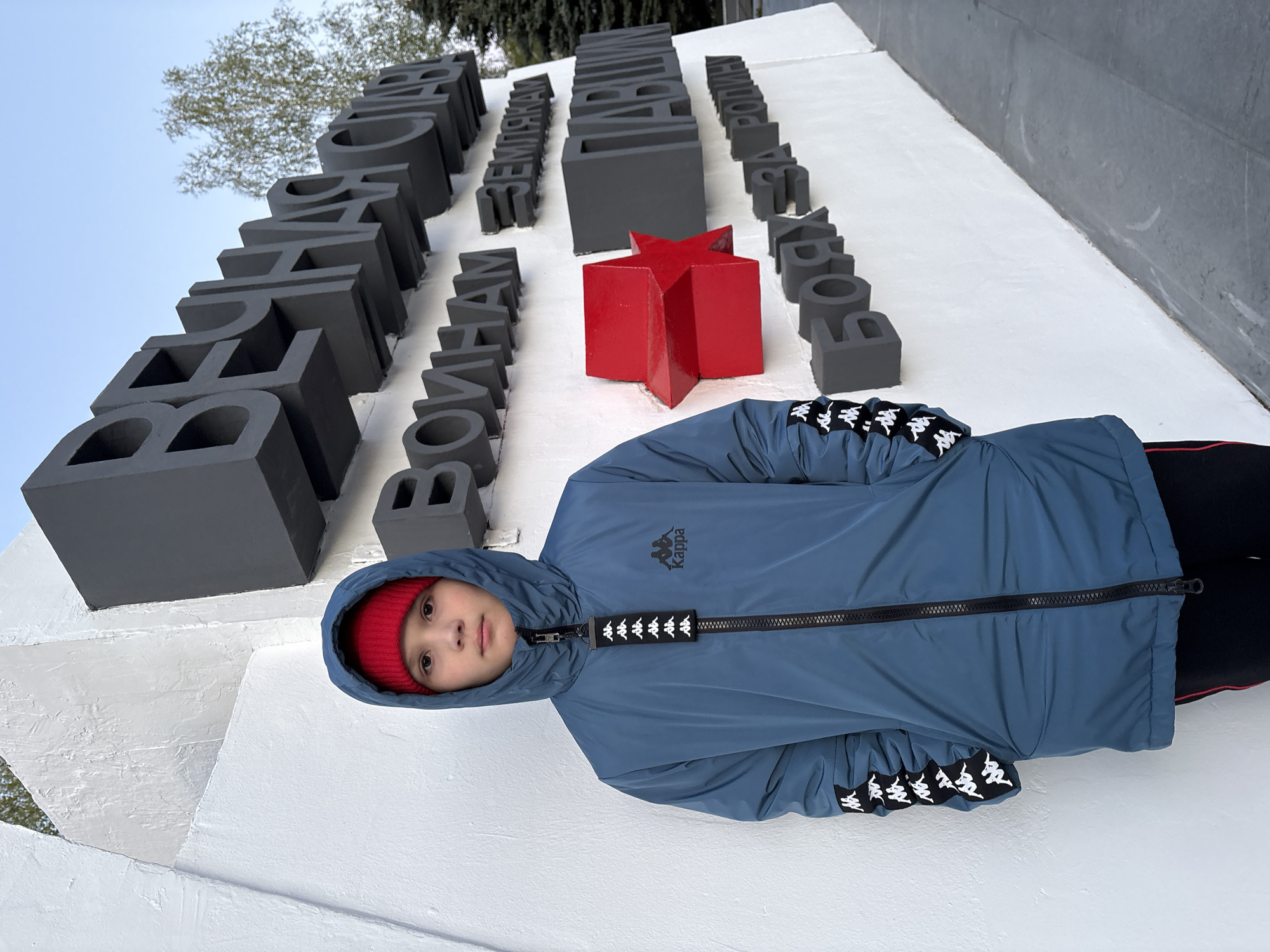 A young child, wearing a red beanie and a blue Kappa jacket, is lying against a white stone monument in Chaikovsky, Russia. The monument features large, dark grey Cyrillic letters and a red star. Part of the inscription "BENGAM SEMLYAKAM GEROYAM RODIN" is visible. The background shows a light blue sky and trees. The photo is taken from a slightly low angle.