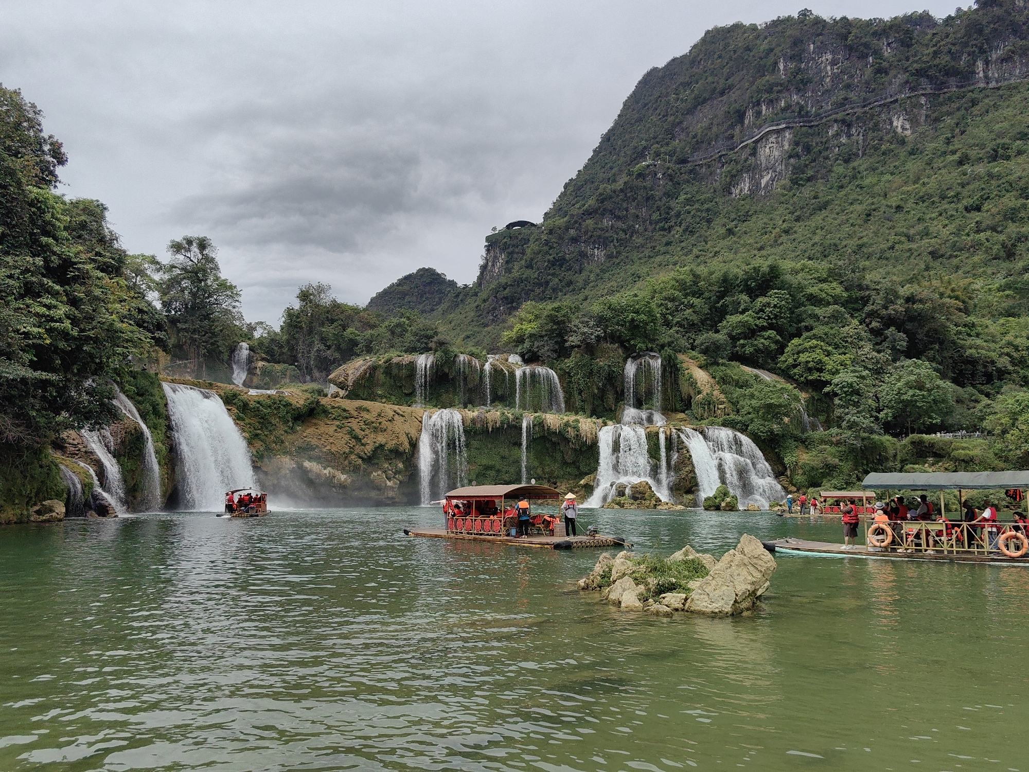 The image depicts the Detian Waterfalls, located in Daxin County, Chongzuo City, Guangxi Zhuang Autonomous Region, China. A wide, tiered waterfall complex cascades into a river. Several rafts are on the water, carrying tourists wearing orange life vests. A person wearing a straw hat stands on one of the rafts. The river is a muted green color. Lush green mountains covered in trees form the backdrop. A pathway is visible on the side of the taller mountain. The sky is overcast and gray. A rock outcropping with vegetation sits in the foreground of the river.