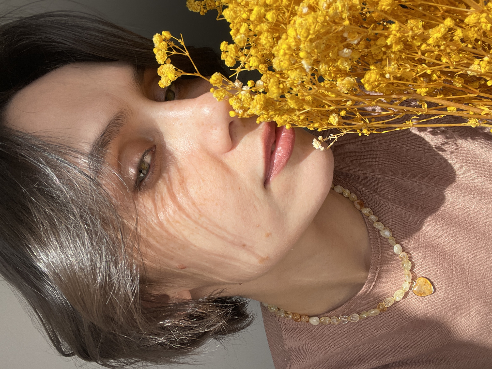 A woman is photographed in a portrait composition, with a cluster of yellow flowers obscuring part of her face. The woman has fair skin, and her features are visible, including her eyes, nose, and lips. Her hair is short and appears dark. She is wearing a light pink top and a necklace with light-colored beads and a heart-shaped pendant. The flowers are positioned near her face, casting shadows across her cheek. The image is well-lit, with natural light highlighting the details of the woman's face, the flowers, and the necklace.
There is no information available in the image to indicate the location is Izhevsk, Russia.