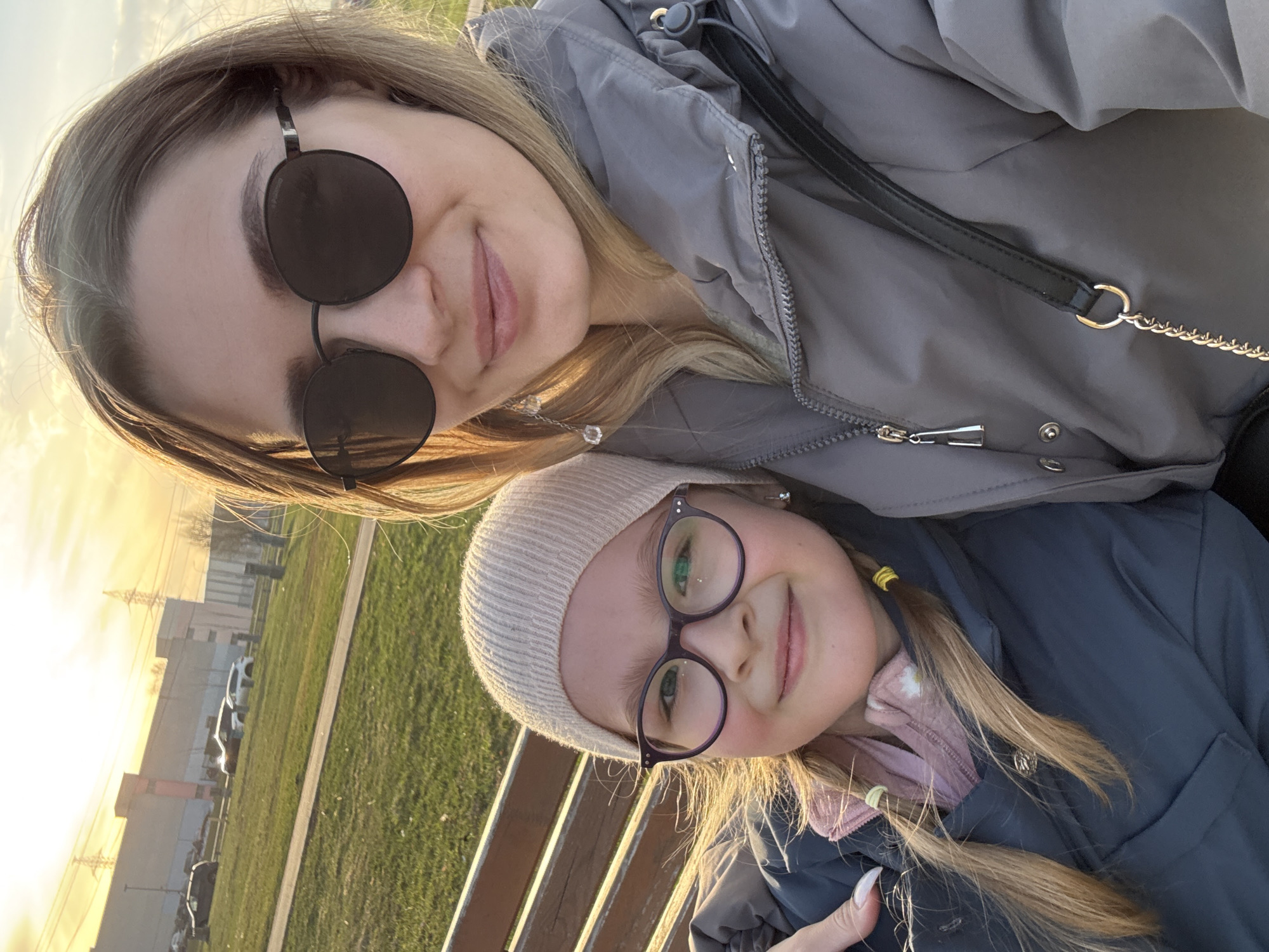 Two females are close together, one adult and one child. The adult is wearing sunglasses and a grey jacket. The child wears a beige hat, glasses, and a dark jacket. The background includes grass, a fence, buildings, cars, and the sky, suggesting an outdoor setting. Given the information provided and the visual characteristics, this image was likely taken in Moscow, Russia.