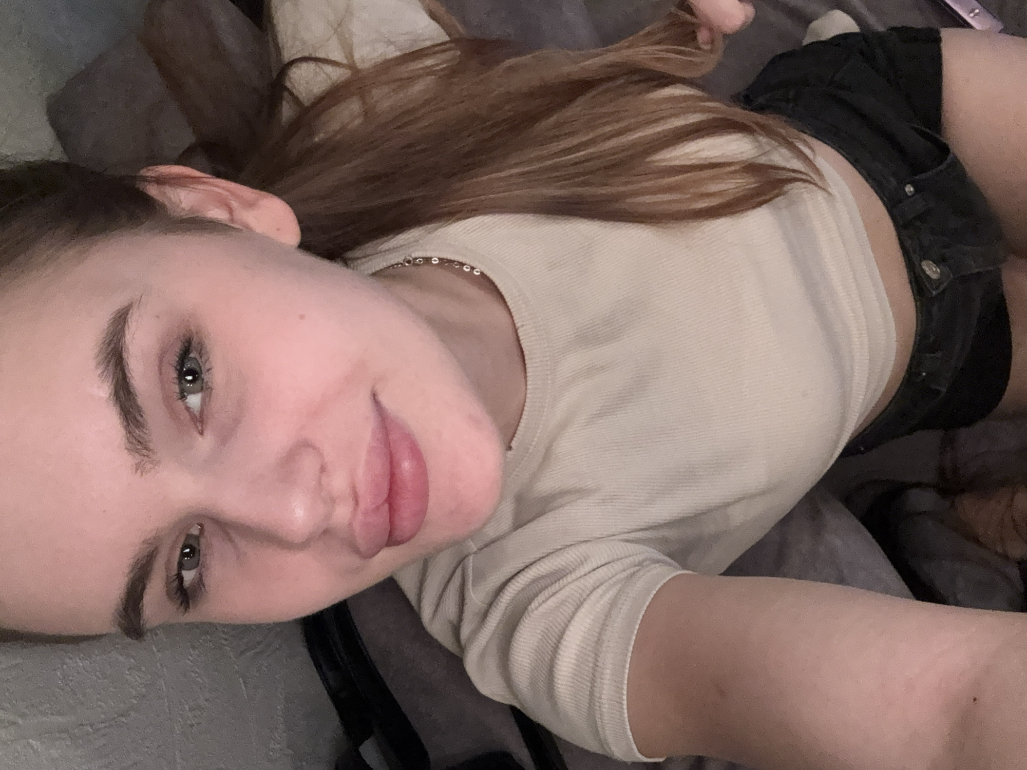 This is a medium-shot, high-angle selfie of a young Caucasian female with fair skin, light brown hair pulled into a ponytail, and light green eyes. She is wearing a beige ribbed crop top and black denim shorts. Her makeup appears minimal, with emphasis on her lips. She is positioned on a bed or couch, indicated by the gray fabric background. There is no discernible location in the city of Dnipro, Ukraine, or any other specific environmental details present in the image. The focus is entirely on the subject's face and upper body.