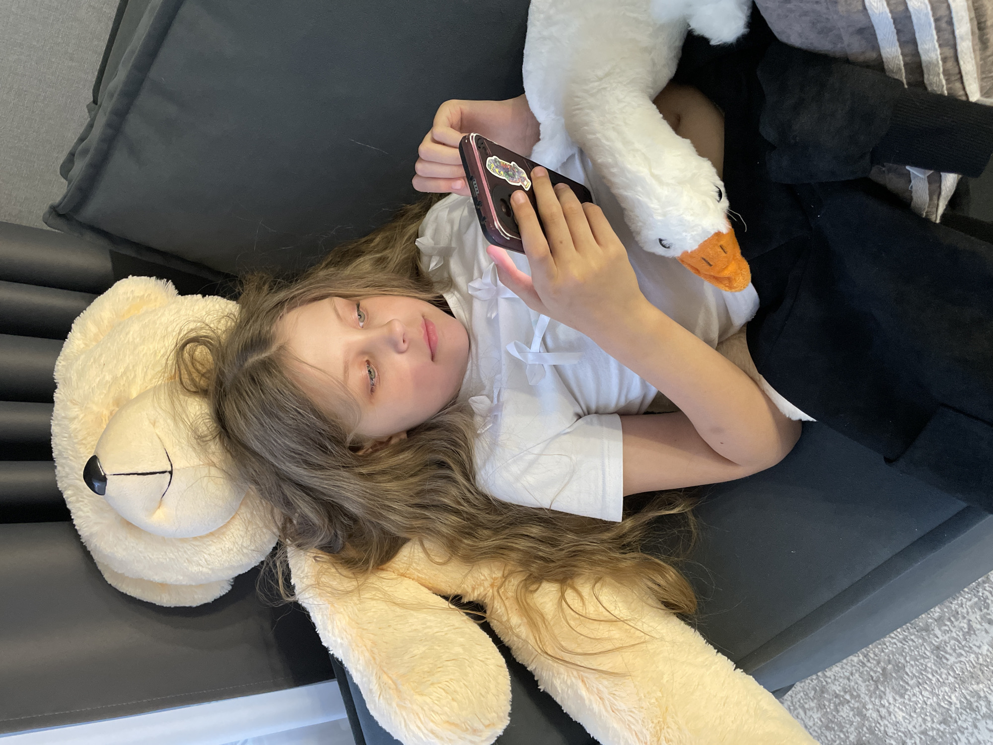 A young girl lies on a dark gray couch. Her head rests on a large, light-yellow teddy bear. She holds a cell phone in her hands, and a stuffed white goose lies next to her. The girl wears a white shirt. The background shows a gray carpet. The scene is likely indoors in a residential setting, although the city of Pervomaysky, Russia, cannot be verified as the location based on the image.