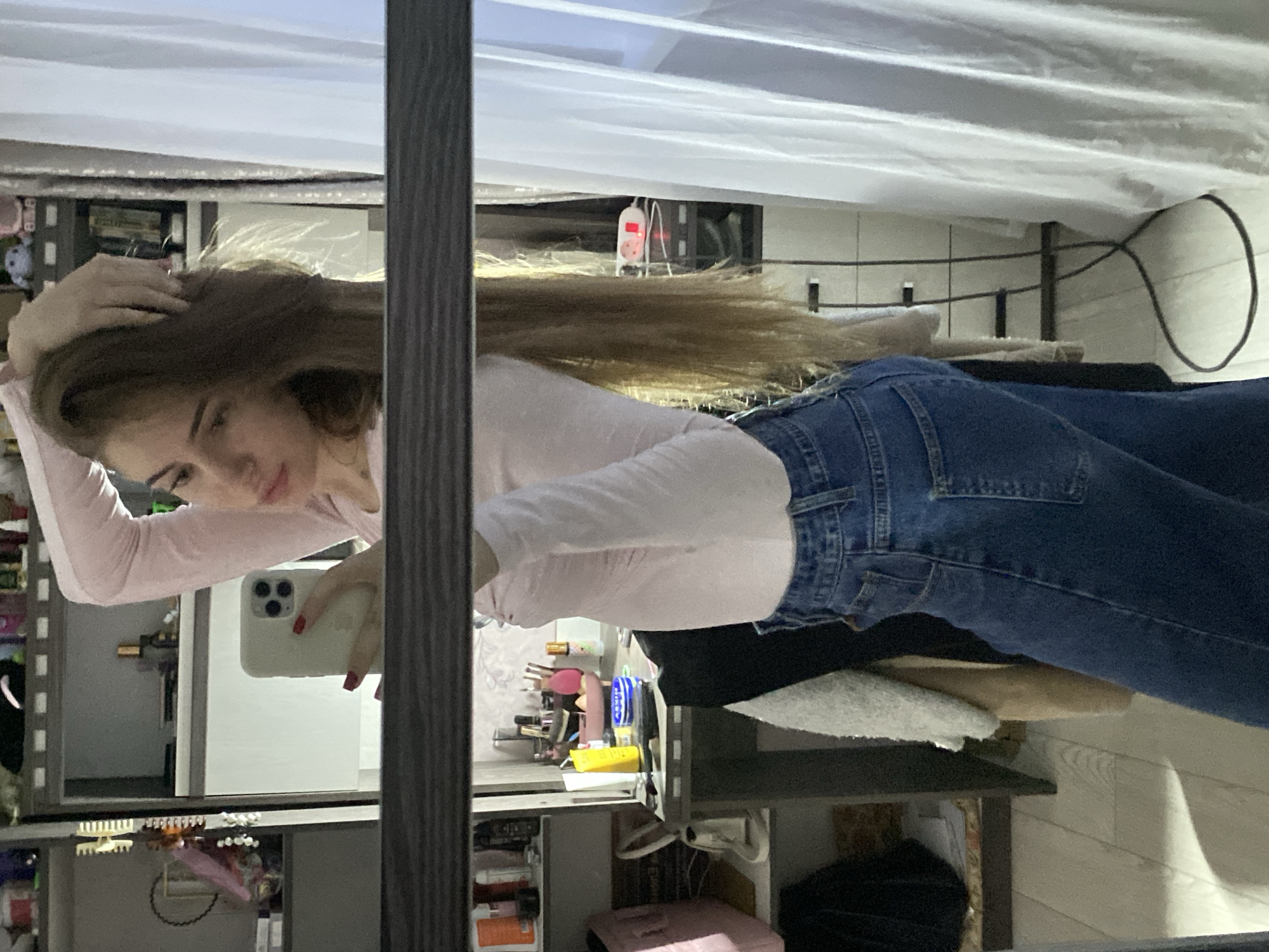 The image is a mirror selfie, rotated 90 degrees clockwise, depicting a young woman in an indoor setting. She has long, light brown hair, which she holds with her left hand, while her right hand holds a light-colored smartphone (an iPhone with three cameras) to take the picture. Her fingernails are painted red.
The woman is wearing a light pink long-sleeved top and blue denim jeans. She looks down towards the phone with a relaxed expression. The background, visible in the mirror's reflection, shows a slightly cluttered room, possibly a bedroom or dressing area. A white, horizontally-striped fabric, which could be a curtain or a bed canopy, dominates the upper part of the reflected background. Below this, various personal items such as makeup brushes, bottles, and other accessories are visible on shelves or a vanity. A small white device with a red digital display is faintly discernible in the distance. A dark, looped cable can also be seen further back. The lighting suggests an indoor artificial environment. No discernible text is visible.