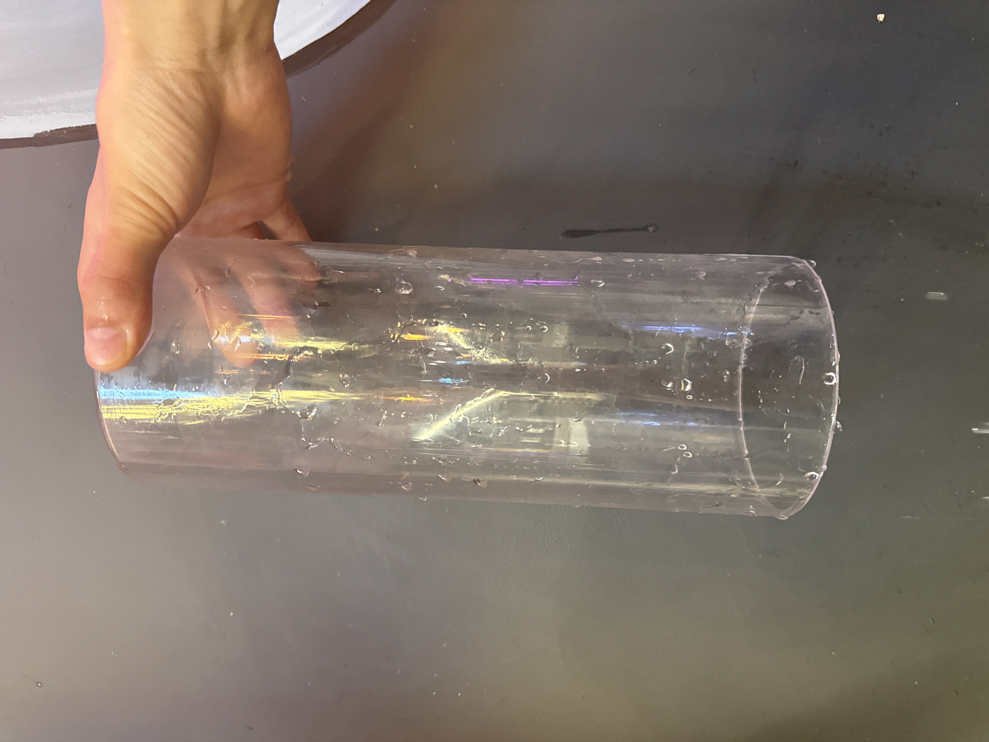 A human hand is holding a clear plastic cylinder over a dark grey surface. The cylinder appears to be slightly wet, with droplets visible on its surface. The surface has some smudges or variations in its color. The location and city cannot be determined from the information available in the image.