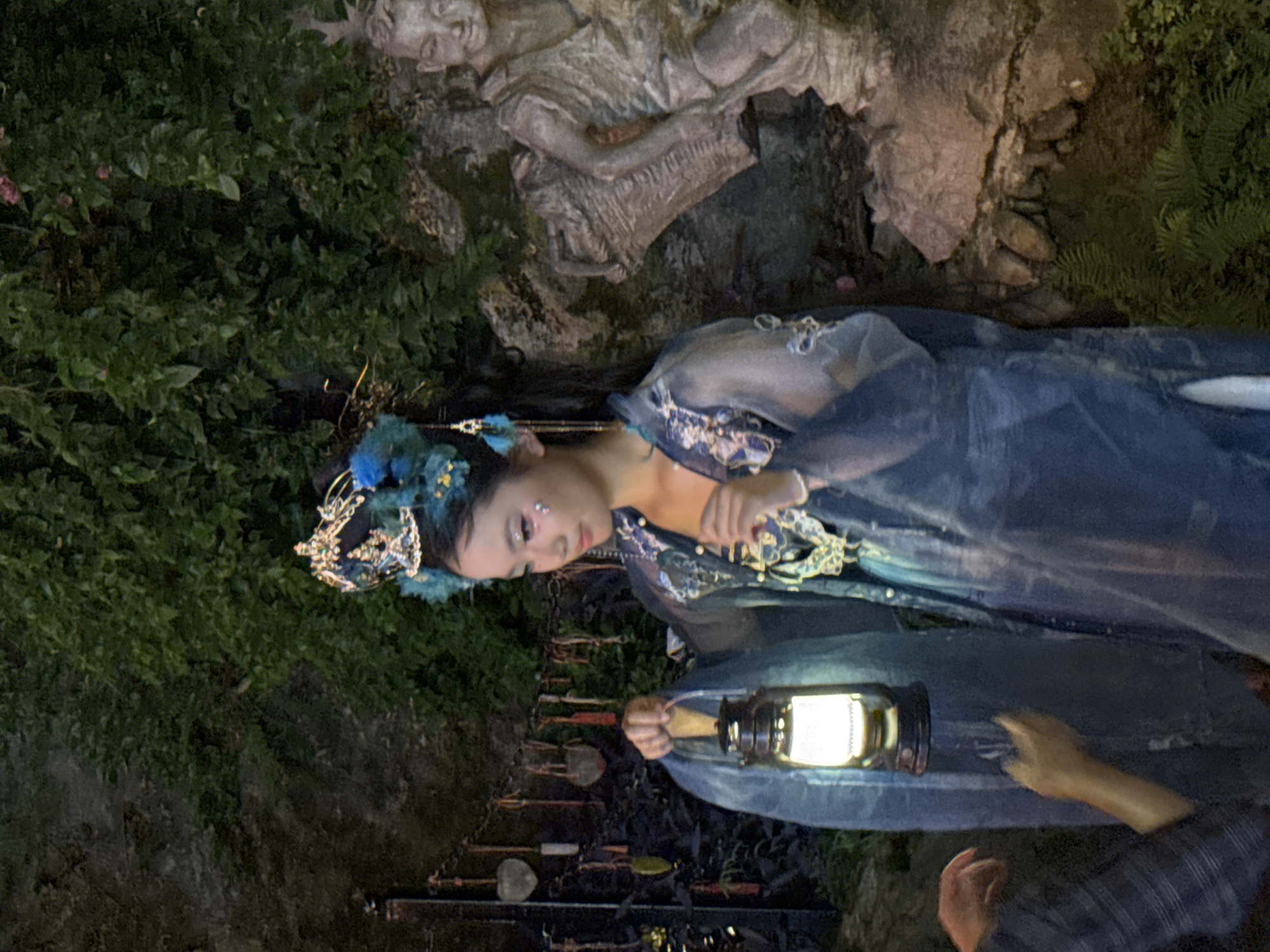 A young East Asian woman, adorned in a blue traditional Chinese outfit and an elaborate headdress featuring blue feathers and gold accents, stands outdoors at night, holding a lit lantern in her left hand. Her face is visible, and she wears glittery makeup on her cheeks. To her right, a stone statue of a figure carrying a basket is partially visible against a backdrop of dense green foliage and rock formations. In the background, chains with hanging heart-shaped ornaments are strung across. Two other individuals are present; one's hands are reaching towards the woman and the lantern from the left side of the frame, and another person's hands are visible near the bottom center of the image. The setting appears to be a garden or a historical site in Yuzhong District, China, possibly illuminated for an event or performance.