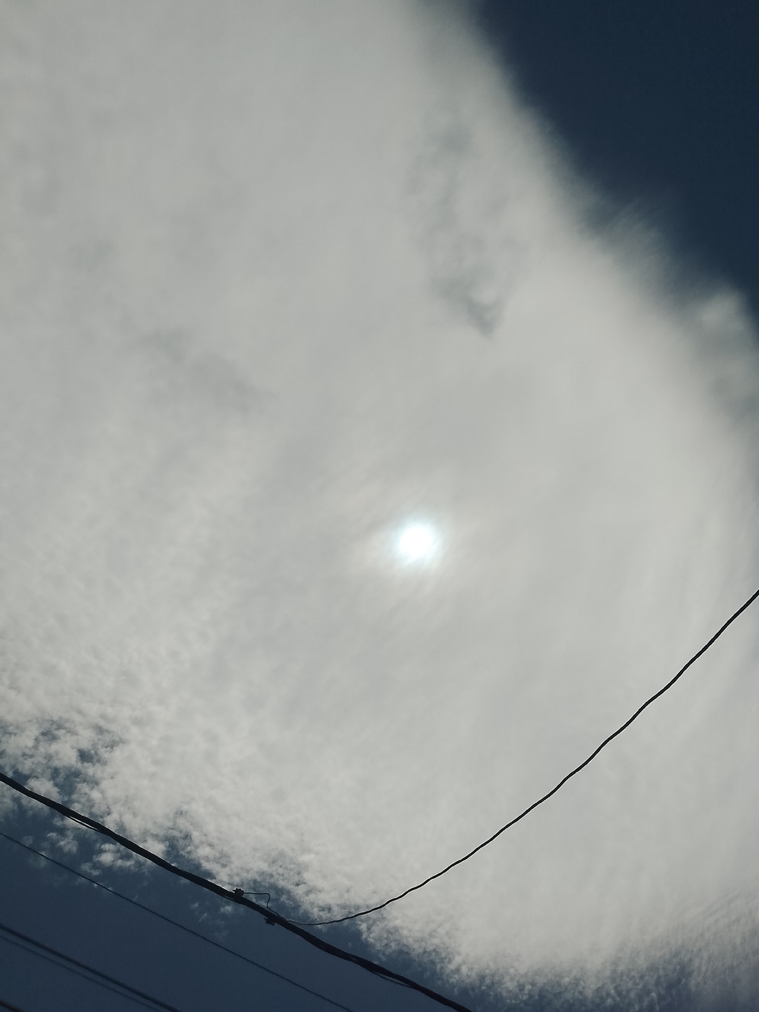 The image depicts an outdoor scene with a bright sun partially obscured by clouds. The sky transitions from a bright, hazy white in the upper half to a darker blue in the lower portion. Thin, dark power lines are visible diagonally across the frame, with one thicker line in the foreground. The overall impression is a slightly overcast day with the sun shining through. The location is specified as Santa Ana, Venezuela, though there are no specific visual cues in the image to confirm this or any urban context.