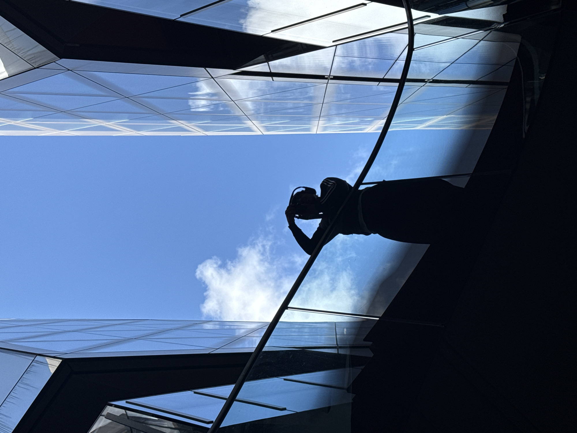 The image is a low-angle shot of a person cleaning windows on a modern building in London, United Kingdom. The person, silhouetted against a bright blue sky with a few clouds, is suspended by ropes and is holding cleaning equipment. The architecture features reflective glass panels arranged in a grid pattern, reflecting the sky and surrounding buildings. The scene is framed by the angular lines of the building's structure.