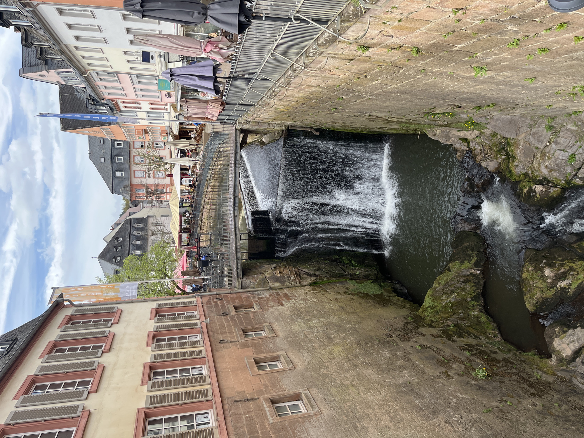 The image shows a waterfall in the city of Saarburg, Germany. The waterfall is located in the middle of the city, surrounded by buildings. The water flows over a series of steps before falling into a pool. On either side of the waterfall are stone walls, with some vegetation growing on the rocks. Along the upper level, on one side, there is a pedestrian area with a metal railing, umbrellas, and outdoor seating, which suggests there are restaurants or cafes nearby. Buildings of various heights and architectural styles line the upper level opposite the stone wall. The sky is visible in the upper left corner, indicating it is daytime. The picture is taken from a high vantage point.
