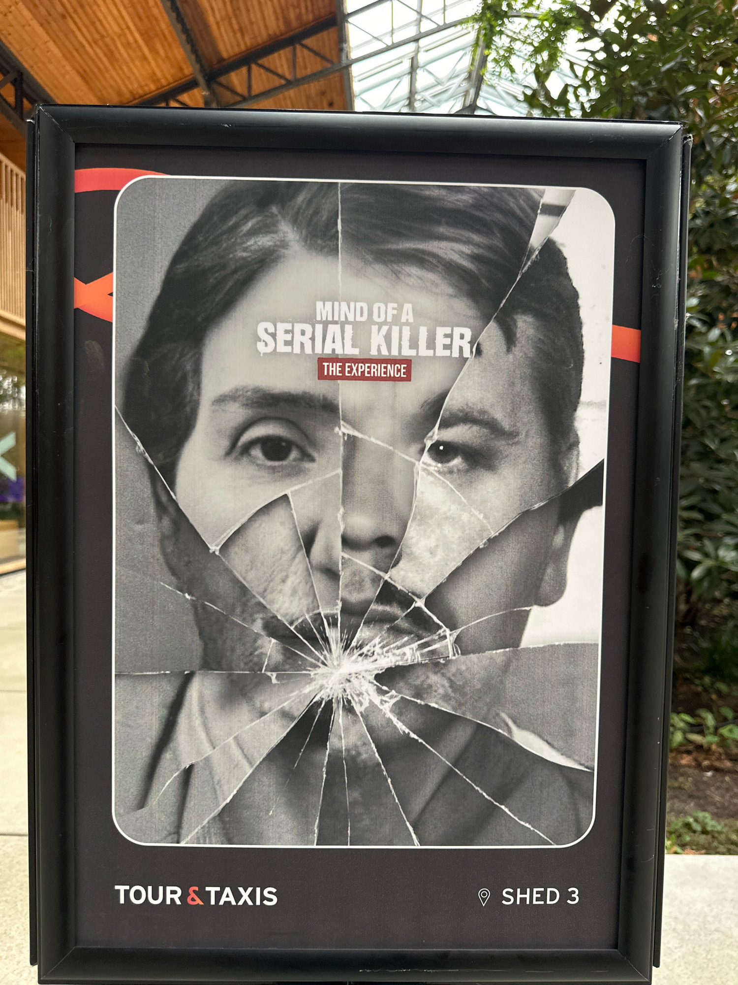 The image displays a framed poster with a cracked glass effect over a black and white photograph of a man's face. Text on the poster reads "MIND OF A SERIAL KILLER" and "THE EXPERIENCE". At the bottom of the poster, there is text that says "TOUR & TAXIS" and "SHED 3". The poster is mounted outdoors, with foliage and a glass-roofed structure visible in the background. The poster appears to be located in the city of Molenbeek-Saint-Jean - Sint-Jans-Molenbeek, Belgium.