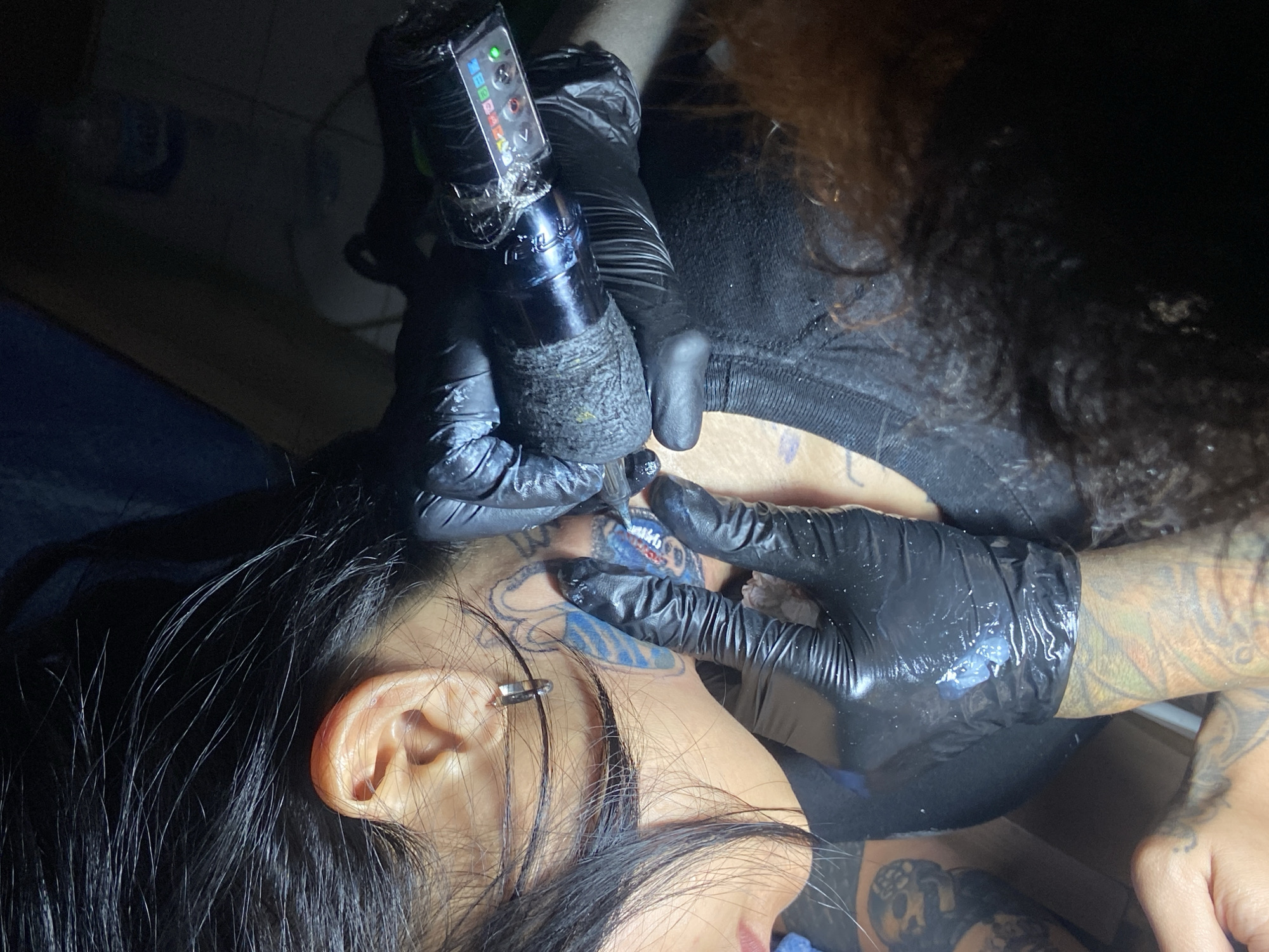 In a dimly lit indoor setting, likely a professional tattoo studio in Kuta, Indonesia, a tattoo artist is actively applying a tattoo to a client's face and neck area. The client, seen from the side, has long dark hair flowing around their head and an ear piercing (a silver cuff or barbell).
The tattoo artist, partially visible with curly brown hair, wearing a black shirt and heavily tattooed forearms, is focused on their work. Both of the artist's hands are covered in black nitrile gloves, which are visibly smudged with tattoo ink and lubricant from the ongoing process. They are operating a modern, pen-style rotary tattoo machine, which is wrapped in clear protective plastic. The machine features a small digital display with illuminated indicator lights (green and red are clearly visible) and control buttons. Text or symbols resembling "ZEBBA" or "ZBX" and directional arrows are discernible on the machine's display.
The tattoo being applied to the client's skin appears to be an intricate design, with existing black outlines and distinct areas of blue shading already present on the skin near the ear and jawline. The overall scene conveys a professional and focused atmosphere during a tattooing session.