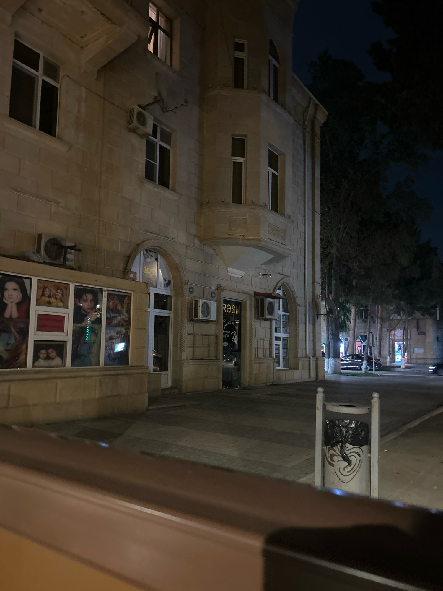 The image shows a nighttime view of a street in Sumgayit, Azerbaijan. A building with light-colored stone facade and multiple windows is prominently featured. Air conditioning units are visible on the exterior of the building. Below the windows, a display of framed pictures, likely posters or artwork, is present. To the right of the building, a street with trees and parked cars is visible, along with streetlights. A trash can with a decorative pattern and a black trash bag is located on the sidewalk. The overall scene suggests a commercial or residential area at night.