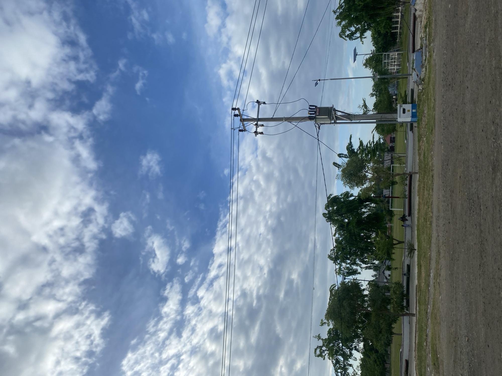 The image shows an outdoor scene, likely in a residential or suburban area in Bireuen, Indonesia. A utility pole with electrical wires dominates the center of the frame, featuring a transformer and insulators. The sky occupies a significant portion of the image, showing a mix of blue sky and white clouds. To the right of the pole are trees, and in the background, some buildings and structures are visible. The right side of the image shows a road or a gravel path.