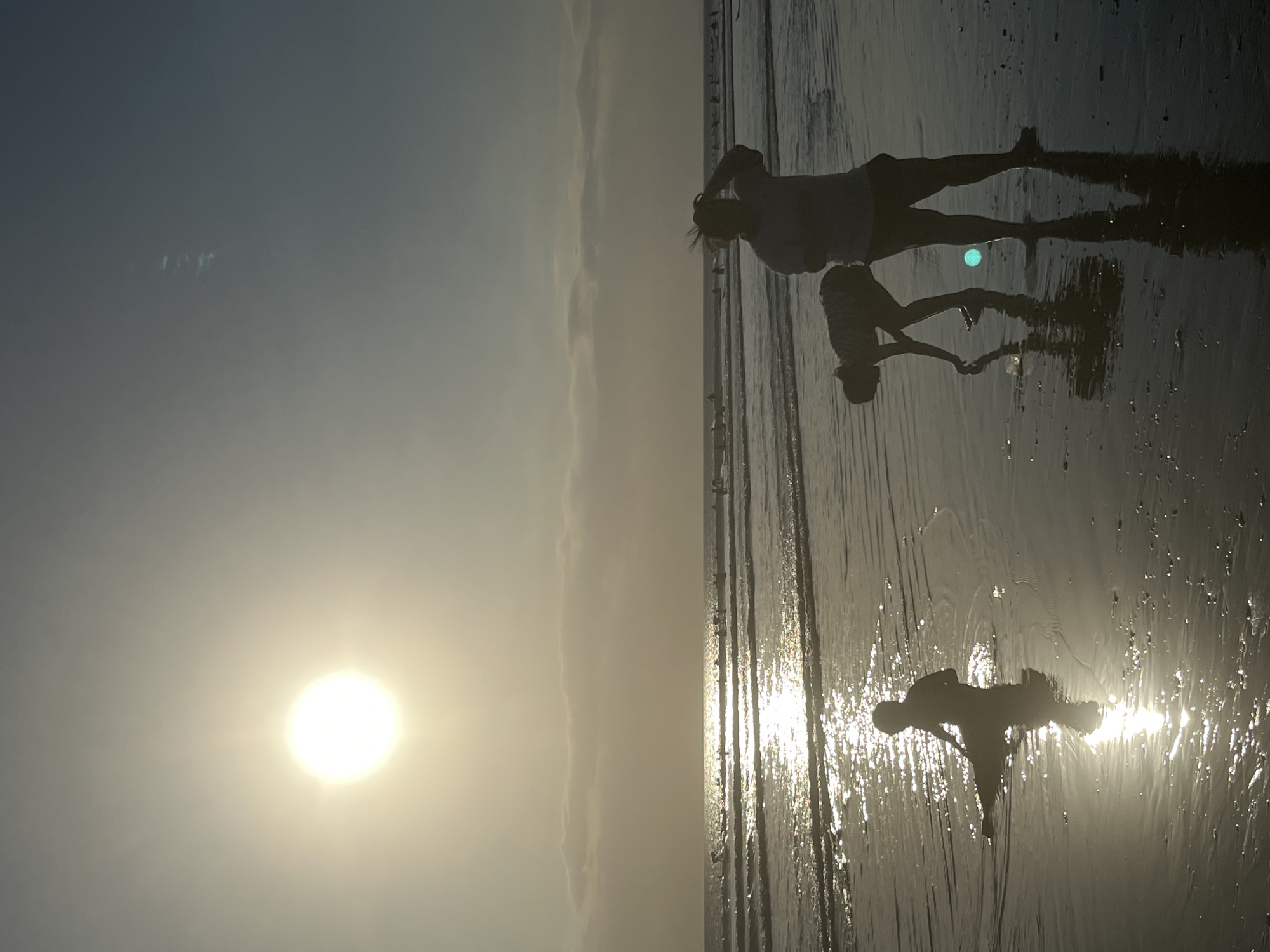 This media file captures a serene sunset scene on a beach, likely in Kuta, Indonesia, as indicated by the location context. The image is oriented vertically, with the ocean and shoreline occupying the right two-thirds and the sky the left third.
In the foreground, silhouetted figures are visible on the wet sand, reflecting the warm light of the setting sun. The first silhouette, closer to the viewer, appears to be a person in a dynamic pose, possibly stretching or dancing, with their reflection clearly visible in the shallow water. Further back, another silhouette depicts two people standing together, their forms also mirrored on the damp surface. The water itself is calm, with subtle ripples and reflections that catch the sunlight.
The sky is a gradient of soft blues and pale yellows, transitioning to a hazy white near the horizon. The sun, a bright, glowing orb, is positioned in the upper left quadrant of the sky, casting a warm, diffused light across the scene. A faint lens flare is visible in the sky, adding to the atmospheric quality of the image. The overall mood is peaceful and contemplative, evoking a sense of tranquility associated with a beach sunset. The wet sand and shallow water suggest a receding tide. There is no visible text in the image.