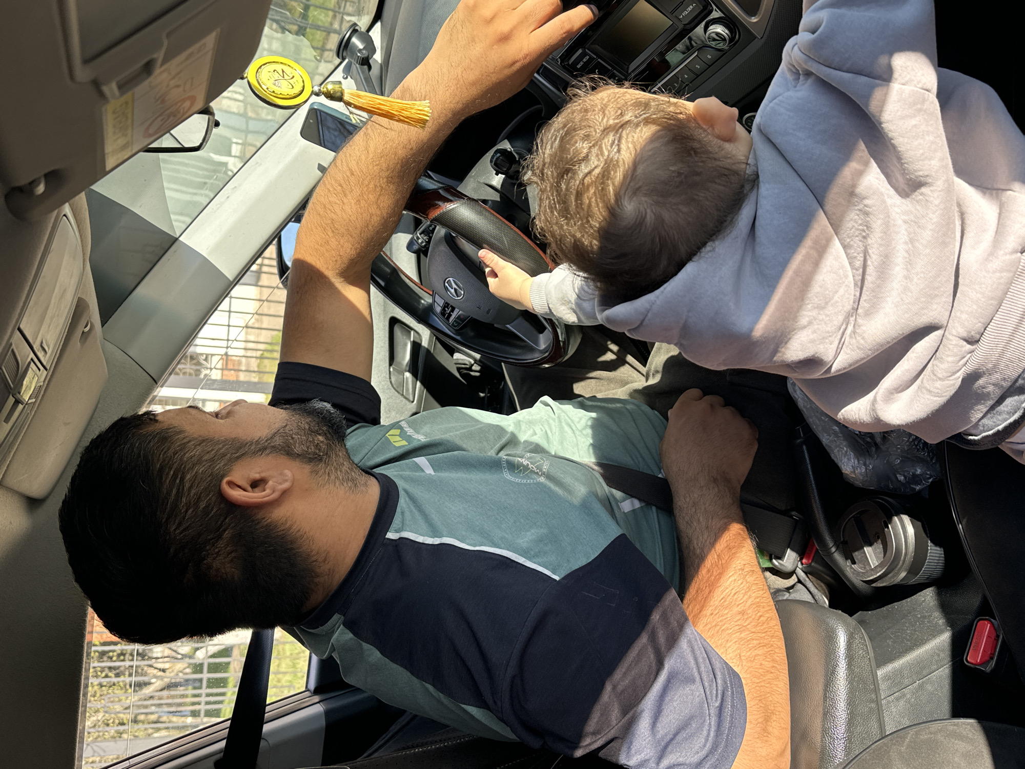 The image captures an overhead view inside a car, likely in Chaikovsky, Russia, during bright daylight. A man with a beard, wearing a green and black short-sleeved shirt, is seated in the driver's seat, with his seatbelt fastened. He is looking out the driver's side window. A young child, dressed in a light grey hooded sweatshirt, is leaning over from the passenger side, reaching their hands towards the steering wheel, appearing to interact with it. The man's left arm is visible, extended towards the dashboard area.
Notable details include strong sunlight casting shadows, indicating a sunny day. The car interior features a dark-rimmed steering wheel, part of the dashboard with controls, and a cup holder in the center console. A yellow circular ornament with a matching yellow tassel hangs from the windshield area. While the specific design on the ornament is not clearly legible, it appears to feature a symbol or stylized letter, possibly a 'M'. Part of an "AIRBAG" warning label is visible on the sun visor. On the man's shirt, a partial logo or text, possibly "MOUNTAIN" or similar, is visible on his right shoulder, along with another indistinct logo on the left. The scene evokes a casual and perhaps playful moment between the man and child in the car.