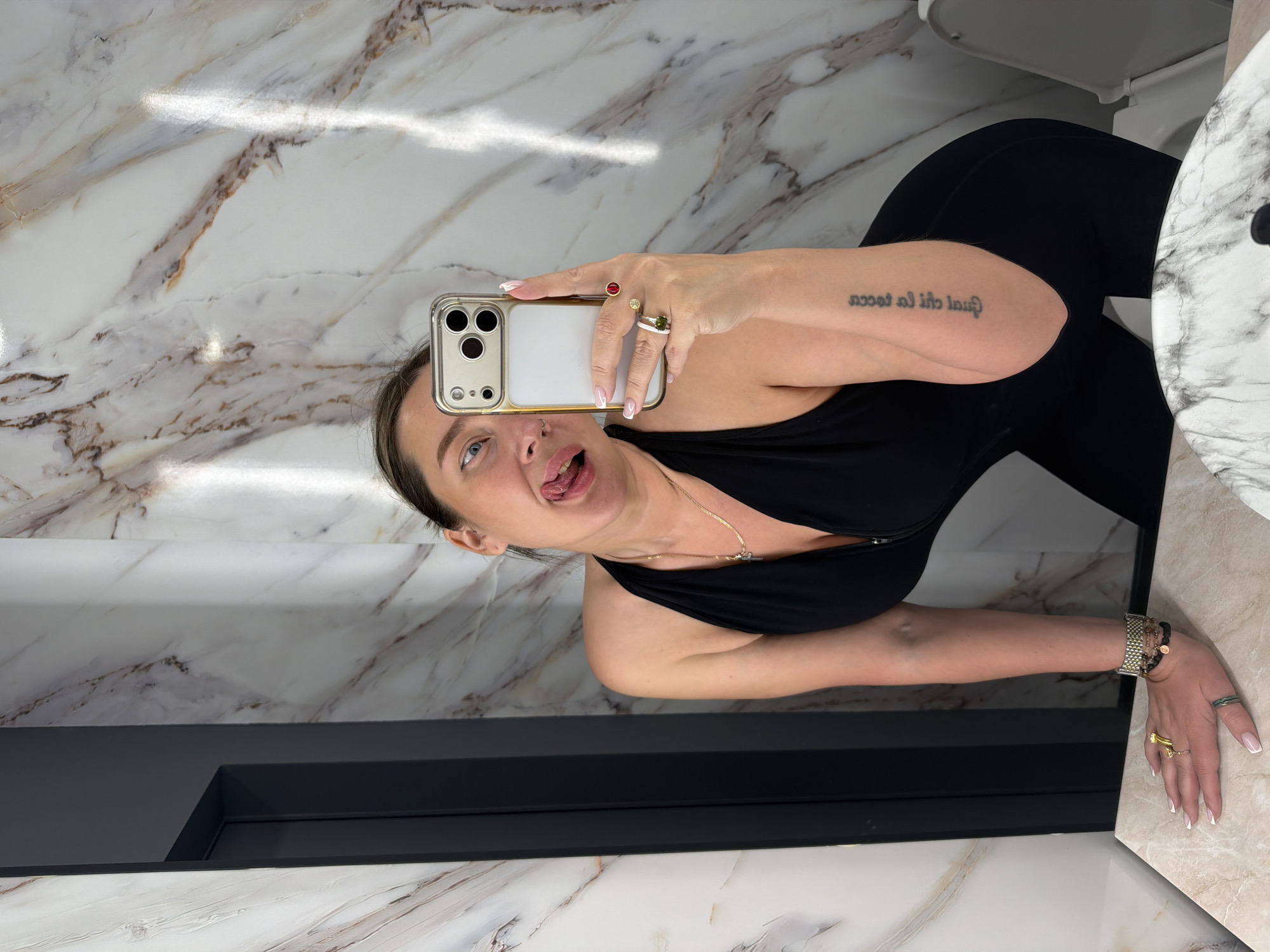 A woman is taking a selfie in a bathroom with marble walls. She is wearing a black top and black leggings. She has a tattoo on her left arm. The bathroom has a toilet, a sink with a marble countertop, and a dark gray vanity. There is no indication of the location in Moscow, Russia.