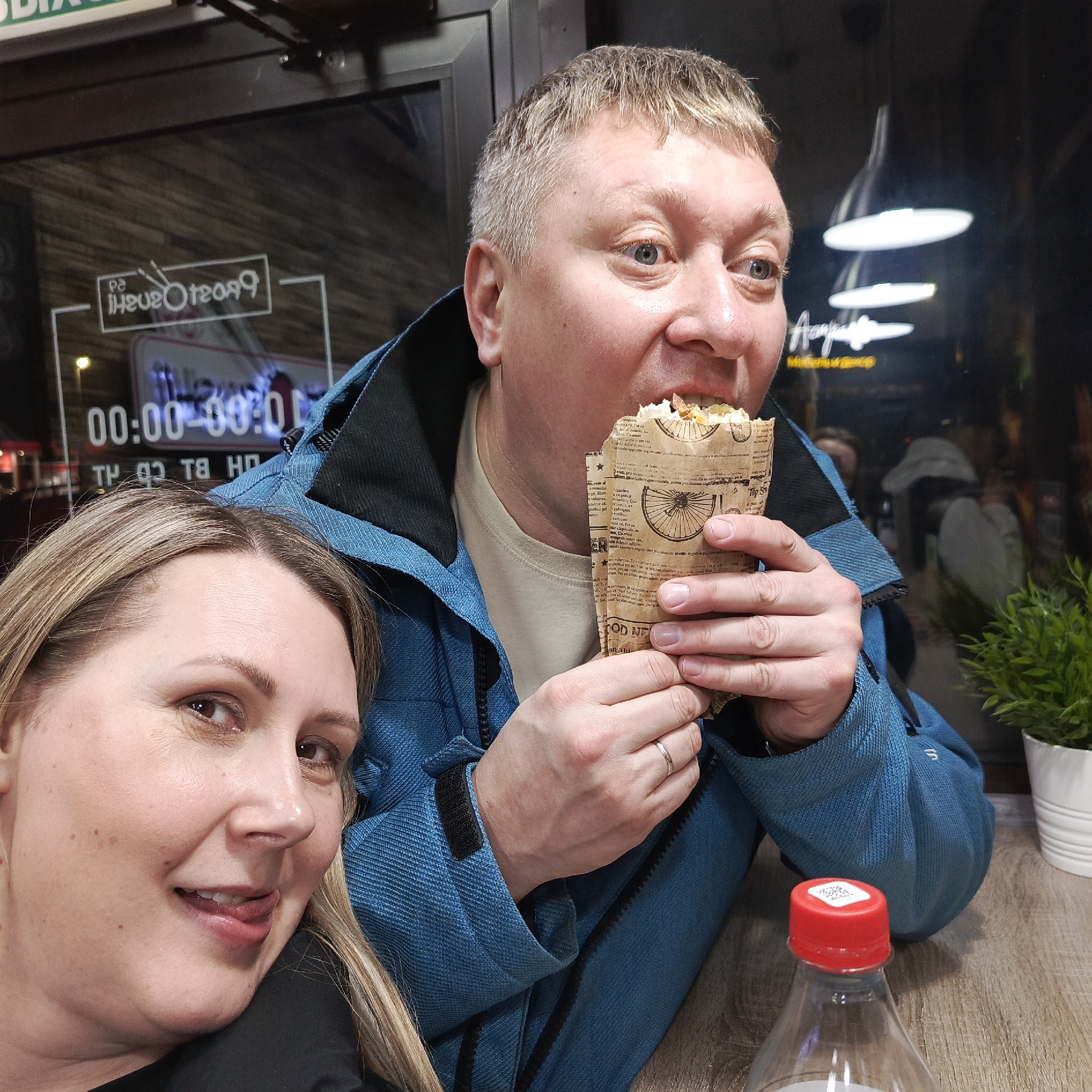 Here is a forensic description of the image:
The image is a close-up shot of a man and a woman sitting indoors at a table, likely a restaurant or cafe. The man, who is positioned slightly to the right, is eating a wrapped sandwich. He is wearing a blue jacket over a beige shirt, and a wedding ring is visible on his left hand. The woman, located to the left, is smiling and sticking out her tongue, wearing a black shirt.
On the table, there is a plastic bottle with a red cap and a small potted plant. Behind them is a glass window with reversed writing, including the words "Sushi" in Russian Cyrillic ("Суши"), and some digital timer display that says "00:00-00:00". Light fixtures are visible in the background through the glass.
Based on the OCR text on the window: "TP 92 T8 HD", it indicates this is a license plate from the Perm Krai region in Russia, which encompasses Chaikovsky. Therefore, it is likely this picture was taken in the city of Chaikovsky, Russia.
