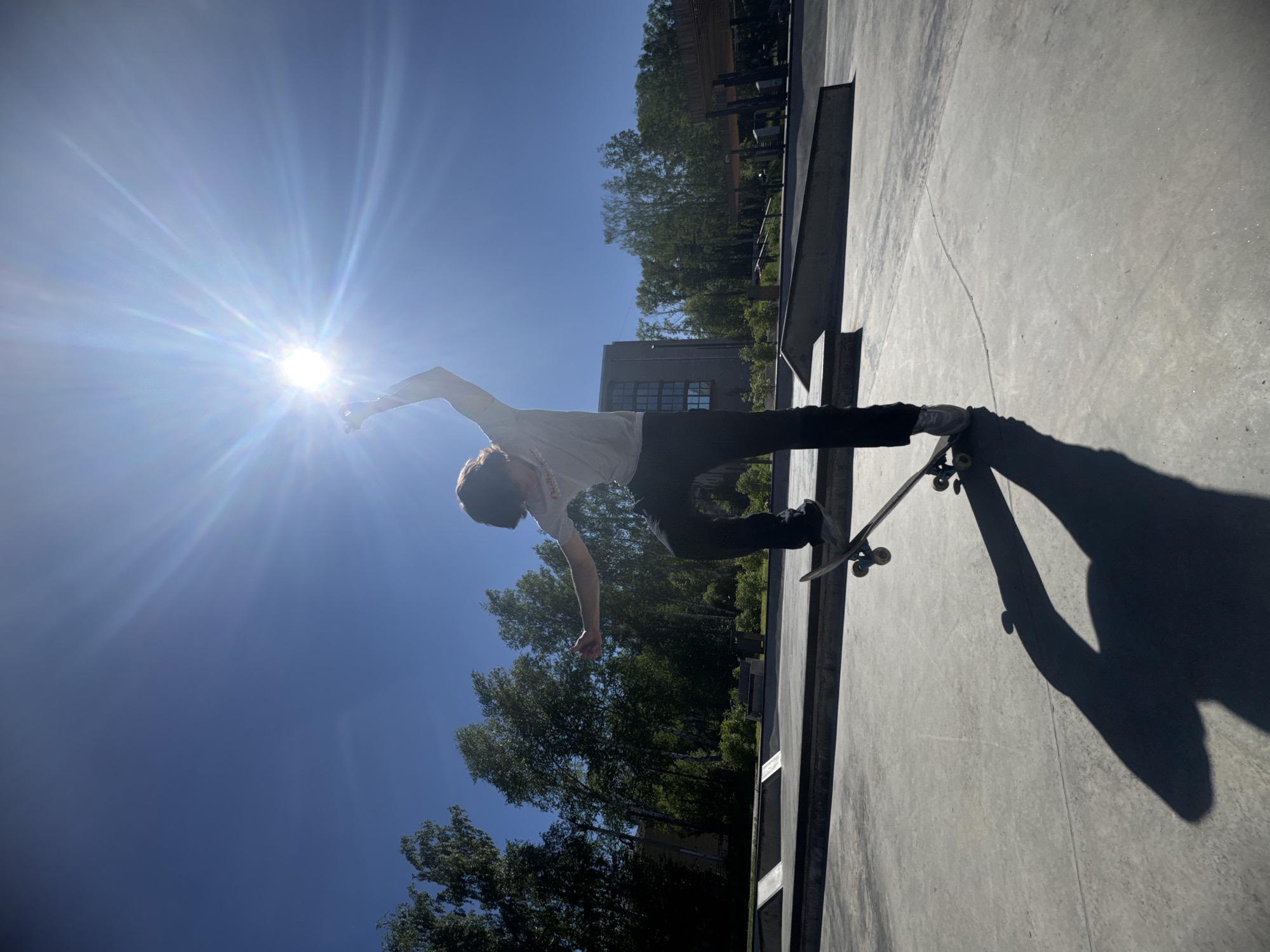In an outdoor skatepark, a person is captured mid-action on a skateboard. The subject, silhouetted against a bright sun, wears a light-colored t-shirt and dark pants. Their left arm is extended upwards, and their right arm is bent at the elbow, with their hand gesturing outward. The skateboarder is positioned over their board, which is angled slightly upwards. The skatepark features concrete ramps and ledges, surrounded by lush green trees and a clear blue sky. The sun is positioned high in the sky, casting a prominent shadow of the skateboarder and their board onto the concrete surface. In the background, a modern building with numerous windows is visible, along with covered seating areas. The scene appears to be set in the city of Divnogorsk, Russia, given the context provided.