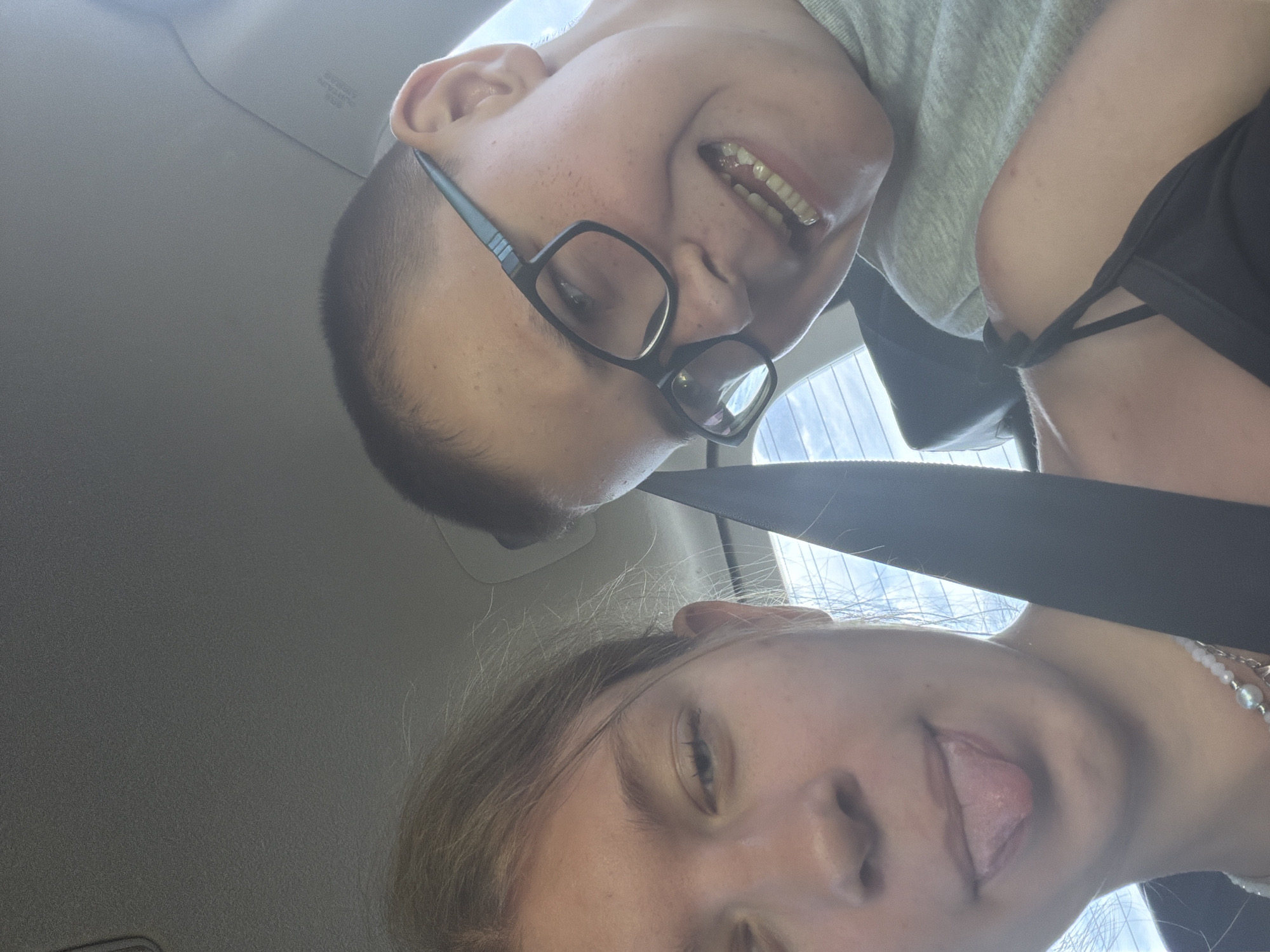 In Ostrovka, Russia, two young individuals are captured in a close-up selfie taken inside a vehicle. The person on the left, a female with light brown hair, is sticking her tongue out. She is wearing a black tank top and a pearl necklace. The person on the right, a male with short dark hair and glasses, is smiling widely, exposing his teeth. He is wearing a grey t-shirt. The background shows the interior of a car, with a seatbelt visible across the frame. The overall impression is a candid and playful moment.