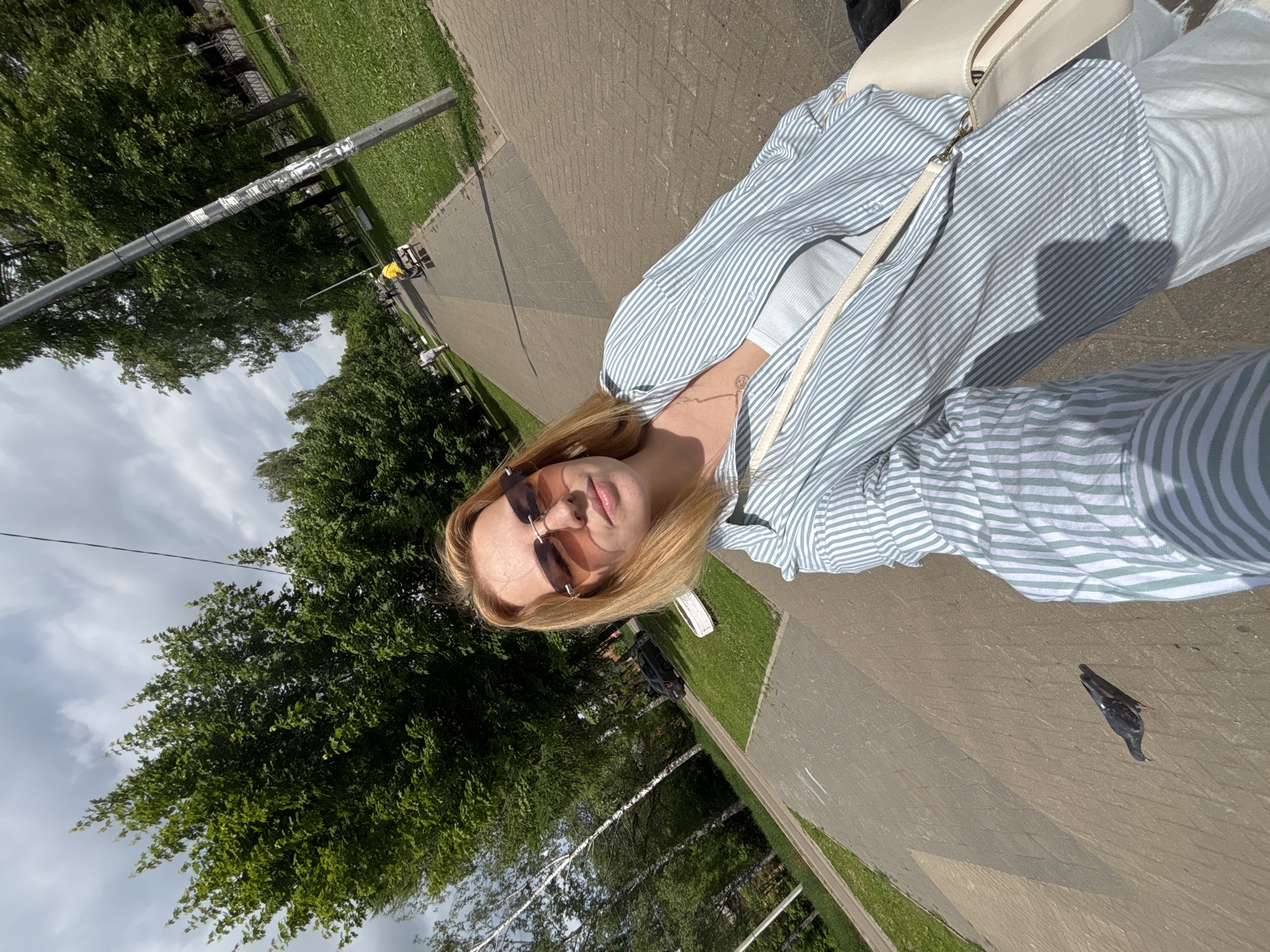 The image captures an outdoor selfie, depicting a young blonde individual standing on a paved path in what appears to be a park or public area, potentially in Podosenikha, Russia. The person wears sunglasses with a reddish tint, a light blue and white striped button-up shirt over a white top, light-colored pants, and carries a white shoulder bag. A subtle necklace with a small pendant adorns their neck. They look directly at the camera with a neutral expression.
The scene is set on a bright, sunny day, likely late morning or afternoon, evidenced by the clear light and shadows, though some clouds are visible in the sky. Lush green trees line the paved pathway, which is constructed from rectangular blocks. Grassy verges run alongside the path. In the background, a tall metal pole with some unclear tape or markings stands vertically. Further along the path, indistinct figures, including one possibly near a cart, are visible in the distance. A pigeon walks calmly on the paved ground in the lower right foreground. No discernible text is visible.