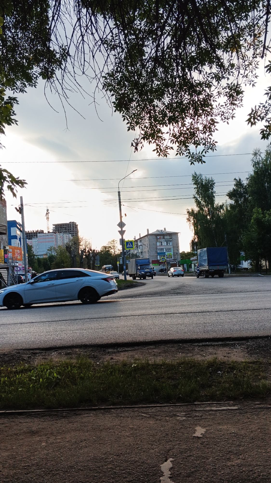The image captures a daytime street scene in Izhevsk, Russia, likely during the warmer months given the foliage. The foreground is dominated by a paved road, with a patch of grass and dirt to the left. The mid-ground features a light blue sedan in motion, a blue truck with a covered trailer, and another white car. Several traffic signs are visible, including pedestrian crossing signs. In the background, apartment buildings and construction sites with cranes suggest an urban environment. Overhead power lines crisscross the sky, which is partly cloudy and bright, indicating late afternoon or early morning light. Trees with dark green leaves partially obscure the sky and buildings from the top of the frame. The overall impression is a typical urban intersection with active traffic.