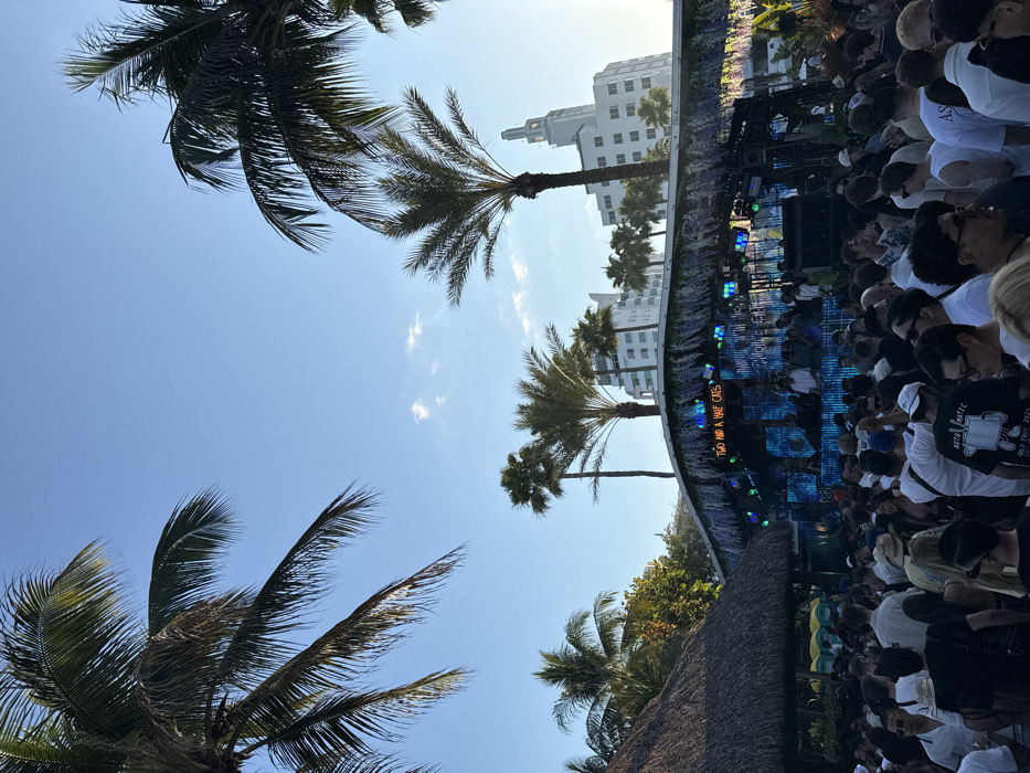 An outdoor music event is depicted under a clear, bright blue sky with sparse clouds, occurring in the city of Miami Beach, Florida, United States of America. In the foreground, a large crowd of approximately two hundred individuals, predominantly adults, is densely packed, facing a central stage structure. Many individuals are wearing light-colored attire, including white t-shirts and hats.
The stage structure features a curved roof decorated with artificial purple and green foliage or lights. A neon sign on the stage reads "TWO AND A HALF CATS." Below this sign, several individuals are visible behind DJ equipment, including what appear to be turntables and mixers, suggesting a performance is underway. The stage is illuminated with blue and green lights. To the left of the stage, a building with a dark thatched roof is partially visible, and further left, two yellow portable toilets are present.
Numerous palm trees of varying heights, including both slender and broader-fronded varieties, are silhouetted against the sky, some framing the upper corners of the image. In the background, multiple white multi-story buildings are visible, including a prominent Art Deco-style structure with a central tower, identifiable as the historic section of the Miami Beach Convention Center, confirming the specific location. The lighting suggests late afternoon, with sunlight coming from the right side of the frame.