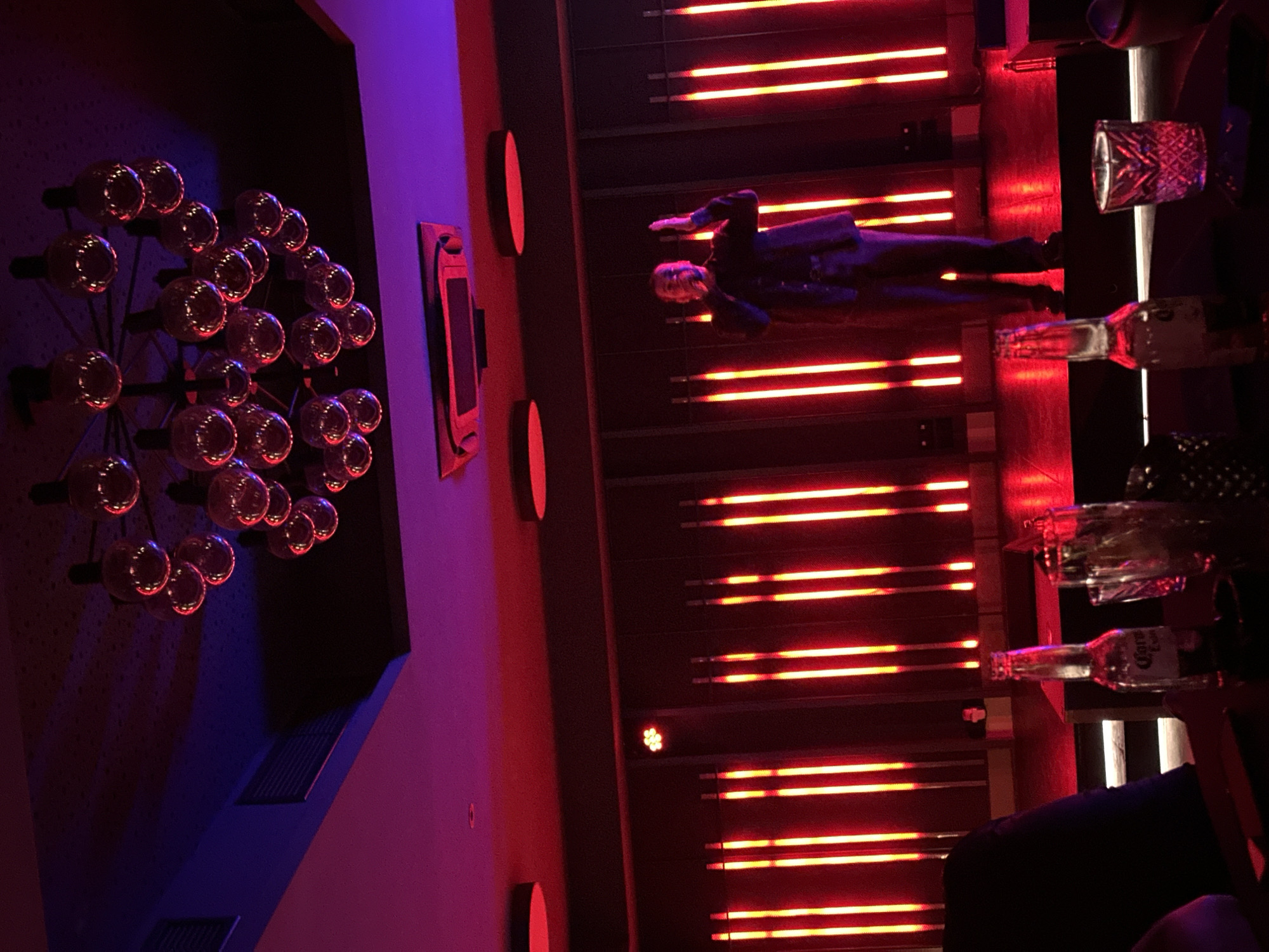 The image depicts a bar interior in Kazanlak, Bulgaria. A person in a dark suit lies on a bar top lit by linear red neon lights. A chandelier consisting of multiple spherical lights is mounted on a dark blue wall. A rectangular object with a display screen is also on the wall. Several drinking glasses and a Corona beer bottle are visible on the bar. The scene is bathed in purple and red light.