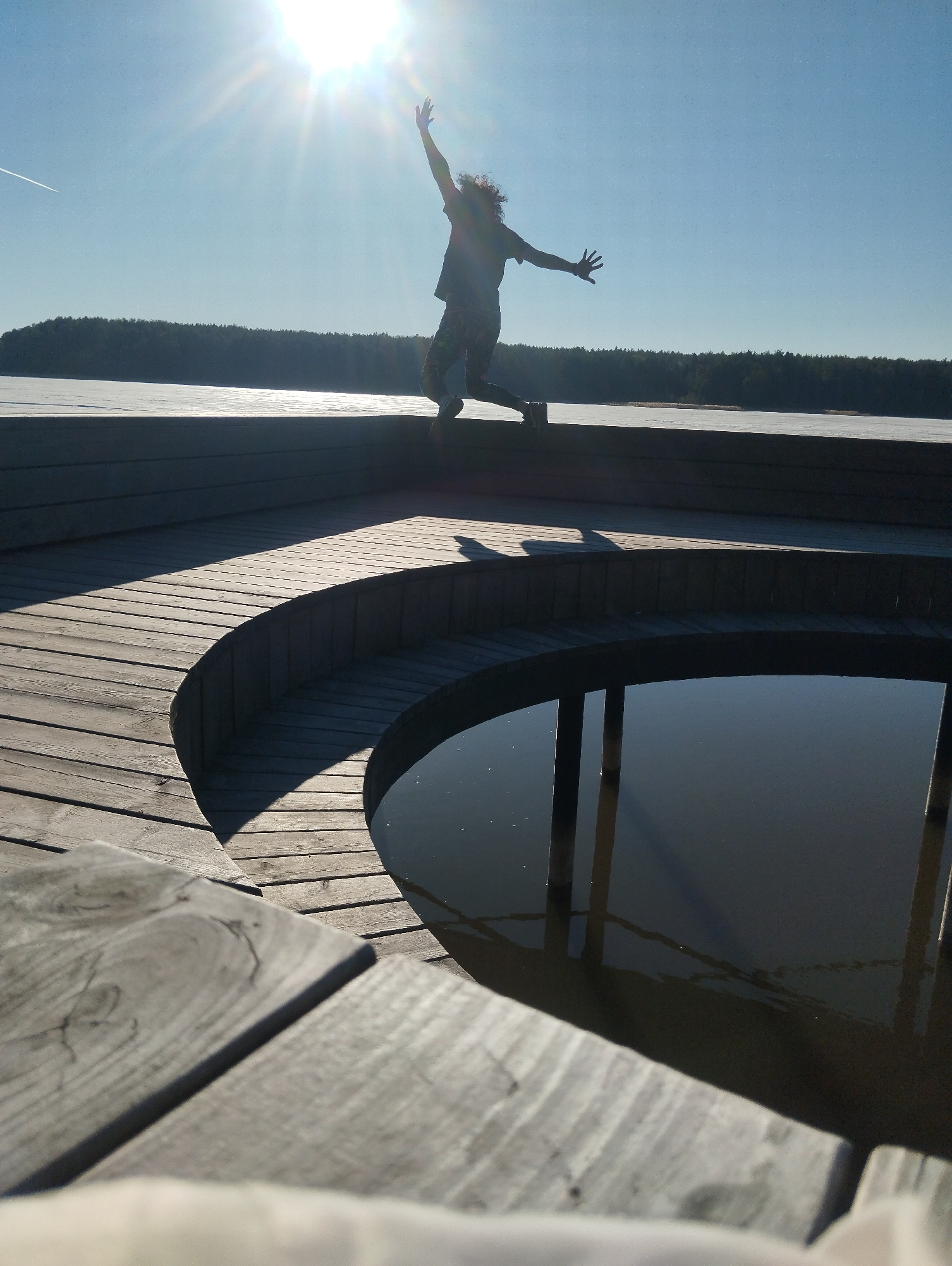 A person is captured mid-air in a jump on a wooden deck, with arms outstretched, against a bright sky and sunshine. The background includes a frozen lake and a treeline. The deck features a circular pool or basin of dark water, reflecting surrounding structures. Based on visual context, this is likely the location of the "Museum of the Votkinsk Factory History" in Votkinsk, Russia.