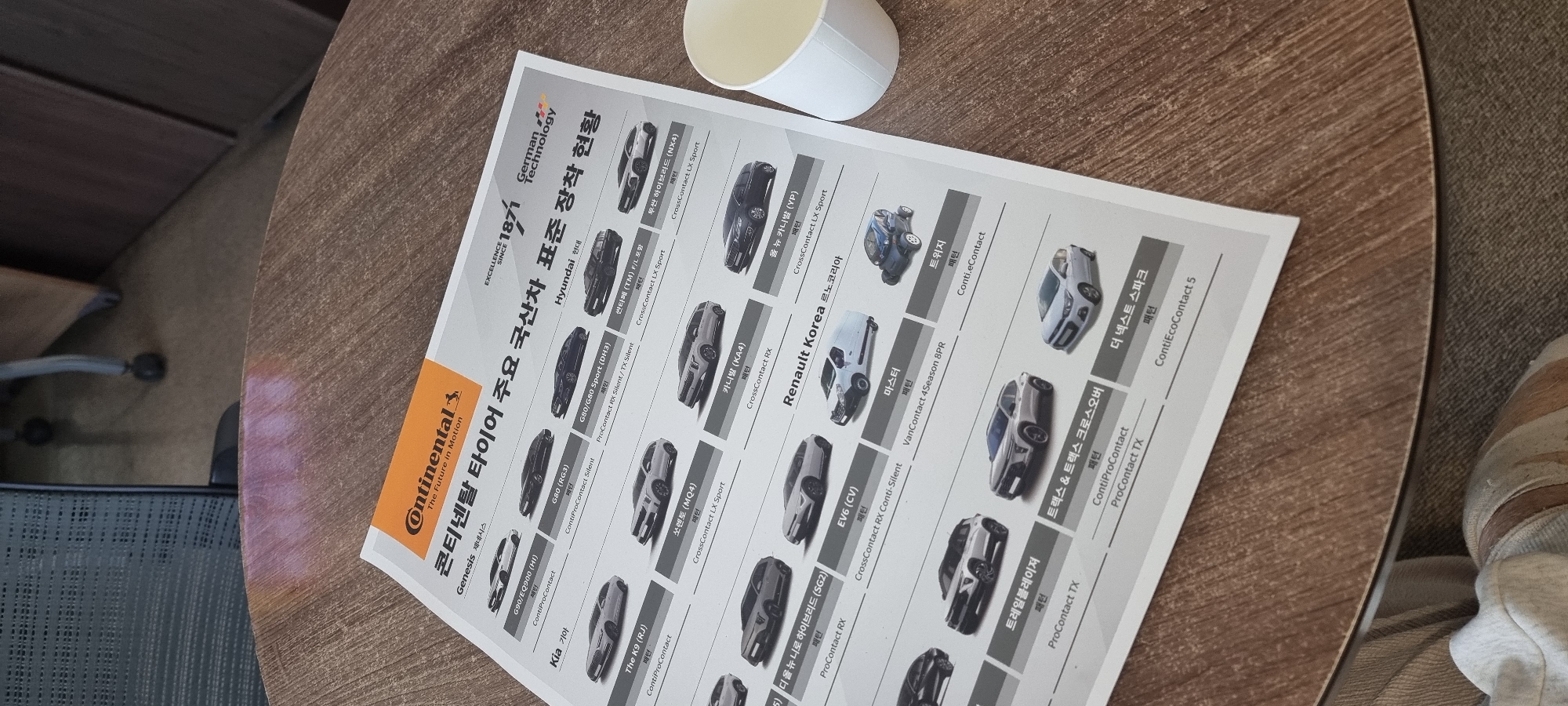 The image shows a Continental Tires advertisement on a table, with a coffee cup resting on top. The advertisement lists various car models and the corresponding Continental tire types recommended for each model. The car brands listed are Genesis, Hyundai, Kia, and Renault Korea. The advertisement also prominently features Continental's logo and the phrase "German Technology".