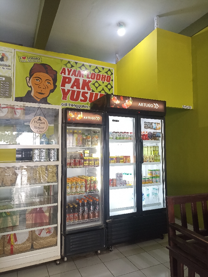 This image captures the interior of a brightly lit eatery, likely a local restaurant or warung in Tulungagung, Indonesia, specializing in "Ayam Lodho." The walls are painted a vivid yellow-green, and the floor is tiled.
A prominent green banner on the back wall features a cartoon-style portrait of an older man wearing a traditional Indonesian head covering. The banner proudly displays "I LOVE LODHO" (with a heart icon), "AYAM LODHO PAK YUSUF," and the phrase "asli trenggalek," indicating the dish's origin from a neighboring regency.
To the left, a tall, glass-fronted display case showcases various packaged snacks, crackers, and what appear to be gift baskets on its lower shelves. A round wooden sign hangs on this case, reading "Pesan & Bayar" (Order & Pay) and "di kasir" (at the cashier). Partially visible next to the display case is a white sign with prices, showing "Rp 7.000" multiple times.
In the center, two large, glass-door commercial refrigerators stand side-by-side, both branded "ARTUGO." They are well-stocked with a variety of cold beverages, including bottled water, juices, and other packaged drinks. One refrigerator also has a "FROST FREE" label. On the far right wall, a small sign for "TOILET" is visible, and part of a wooden bench or chair can be seen in the foreground, suggesting a dining area. The overall scene suggests a clean and inviting establishment.